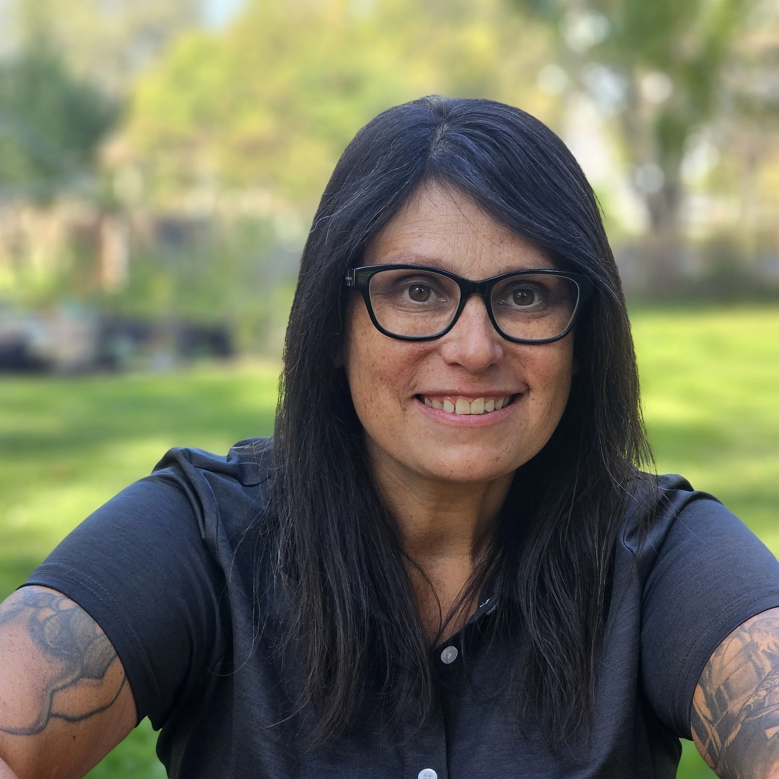 A smiling woman with black hair, glasses, and tattoos on her arms taking a selfie outdoors with green trees and a blurred background.