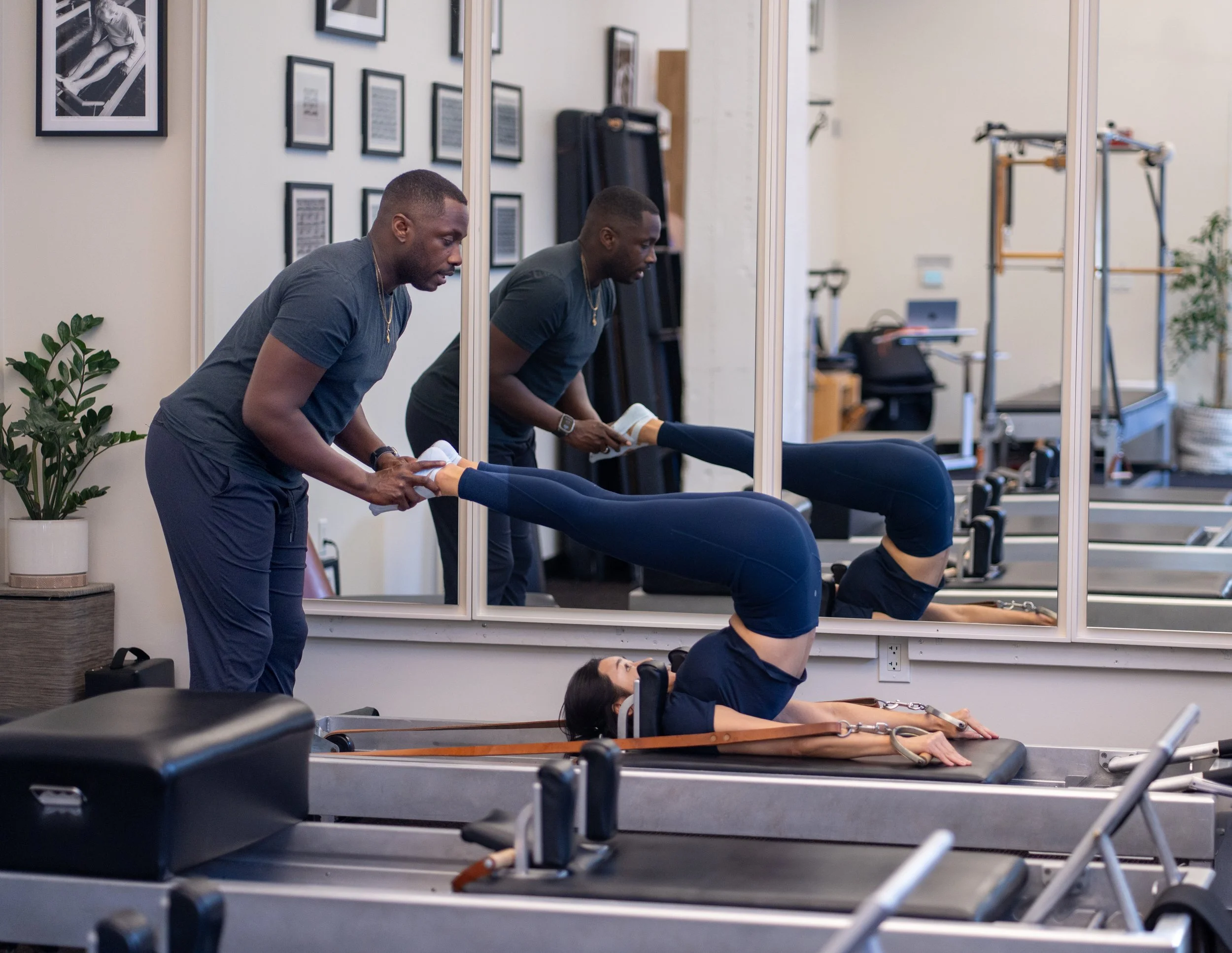 A personal trainer assisting a woman with a workout on the reformer in a fitness studio with framed pictures on the wall, a mirror, and pilates equipment.