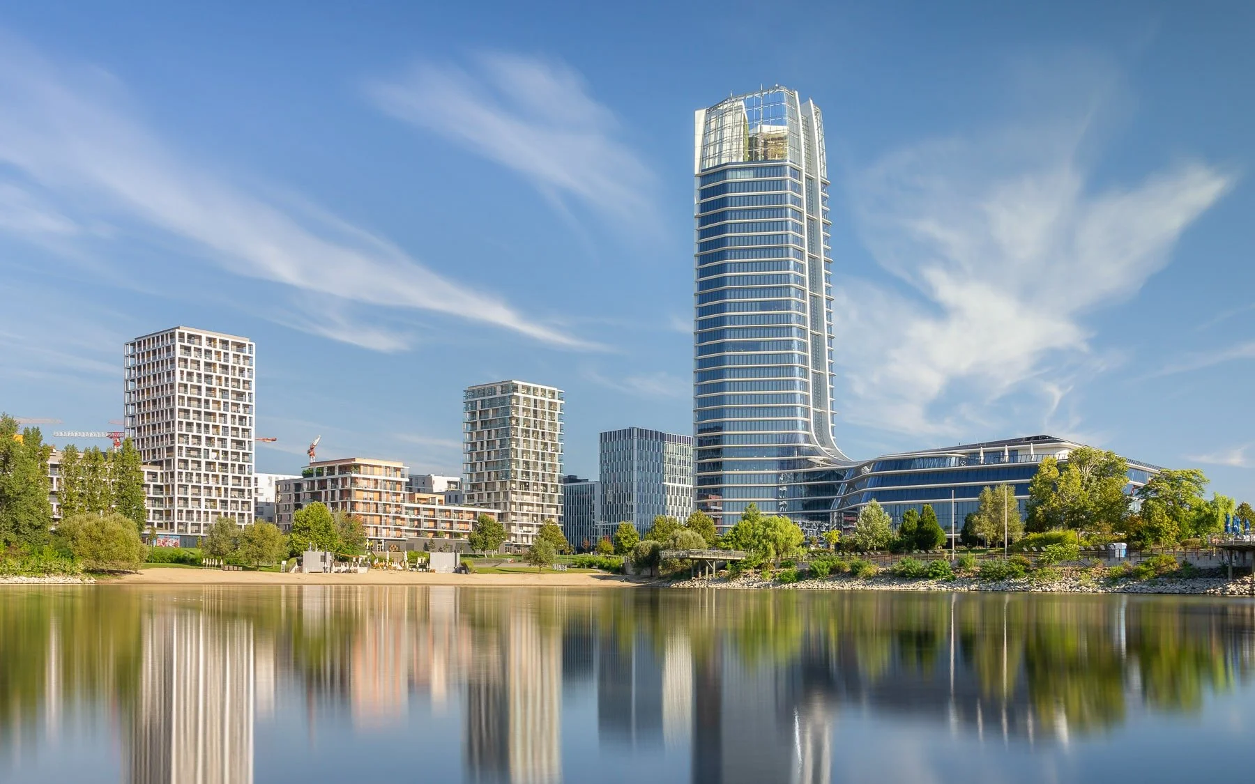 Budapest, Hungary: MOL Campus tower, a modern skyscraper with a striking curved design, reflected in the Danube River, creating a visually interesting composition. Other contemporary buildings and green spaces are visible in the background.