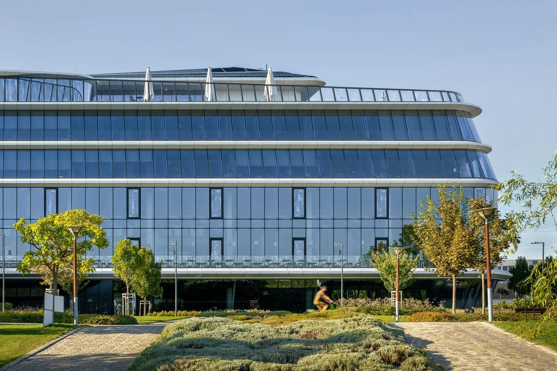 Budapest architecture: Contemporary MOL Campus building showcasing its sleek, curved design, reflective glass surface, and rooftop elements. Green spaces, trees, and a cyclist are visible in the foreground.