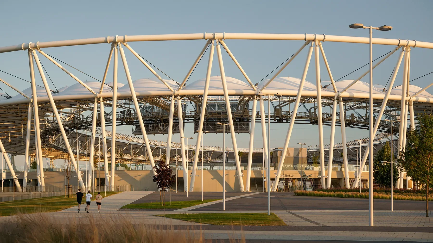 Wide exterior shot of Budapest Athletics Stadium showing full structure, surrounding park, shadow play, and people running