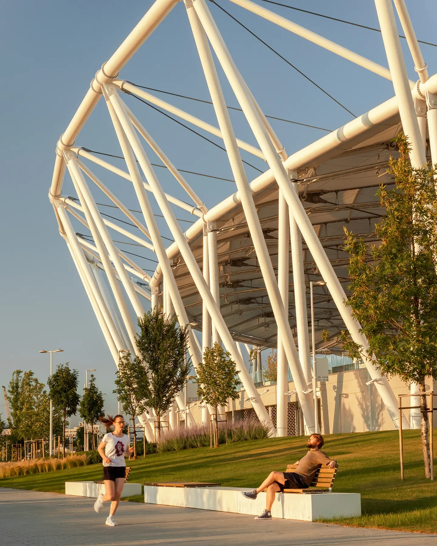 Exterior side view of Budapest Athletics Stadium showing metal structures, runner, bench, and greenery near river at sunset