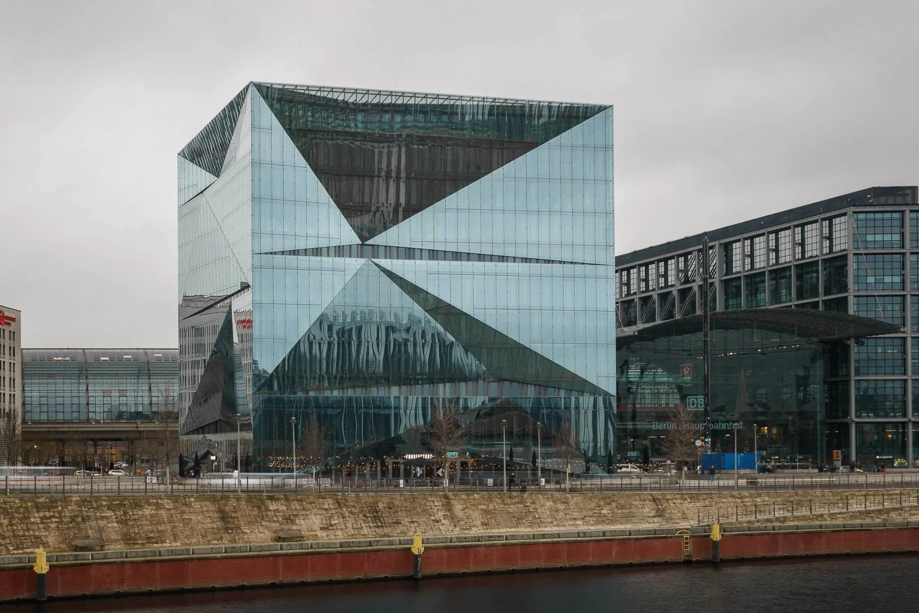 Geometric elegance of the 3XN Cube, Berlin. Riverfront view showcases its unique architectural design under a cloudy sky