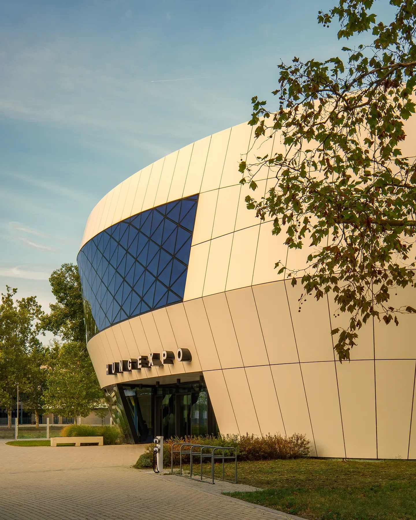 Right-side view of Hungexpo F1 Reception Building in Budapest, glass curves glowing under dark blue sky with trees in foreground