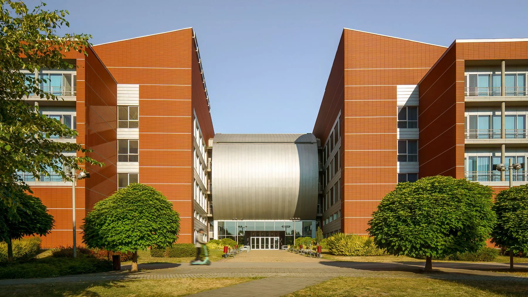 Back of BME Q Building at Infopark Budapest fully sunlit with person for scale, showing metal roof and structural design
