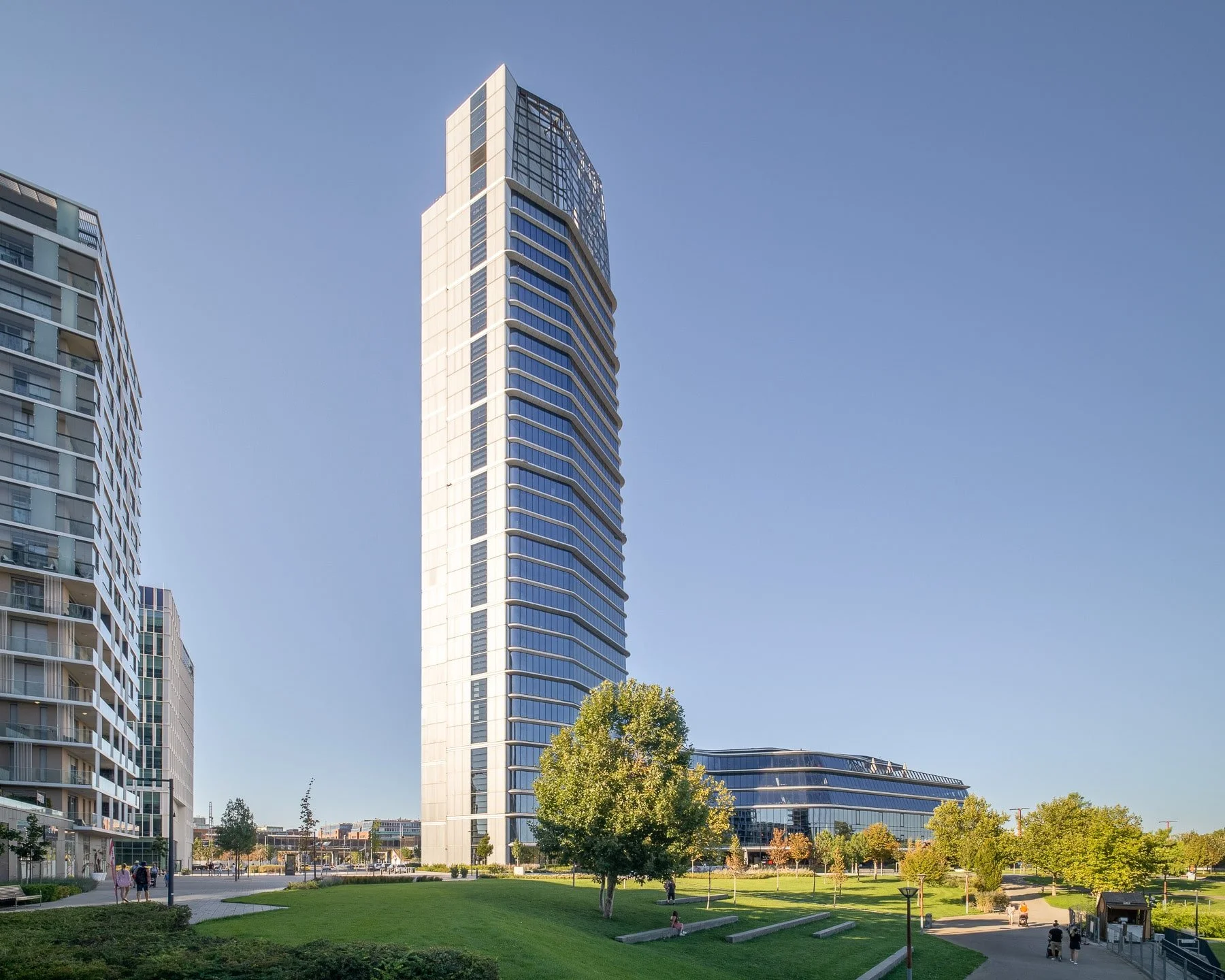 Budapest architecture: Contemporary MOL Campus building showcasing its sleek, curved design, reflective glass surface, and exposed framework. Green spaces, trees, and pedestrians add scale to the scene, with a clear blue sky in the background.