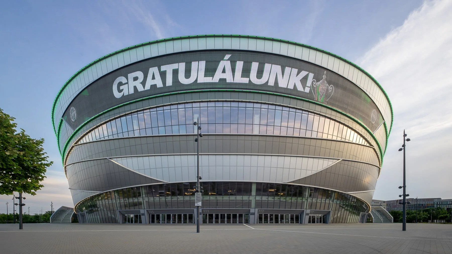 Budapest architecture: MVM Dome featuring its distinctive curved shape, glass facade, and the "GRATULÁLUNK!" message.  Early evening view with clear skies.