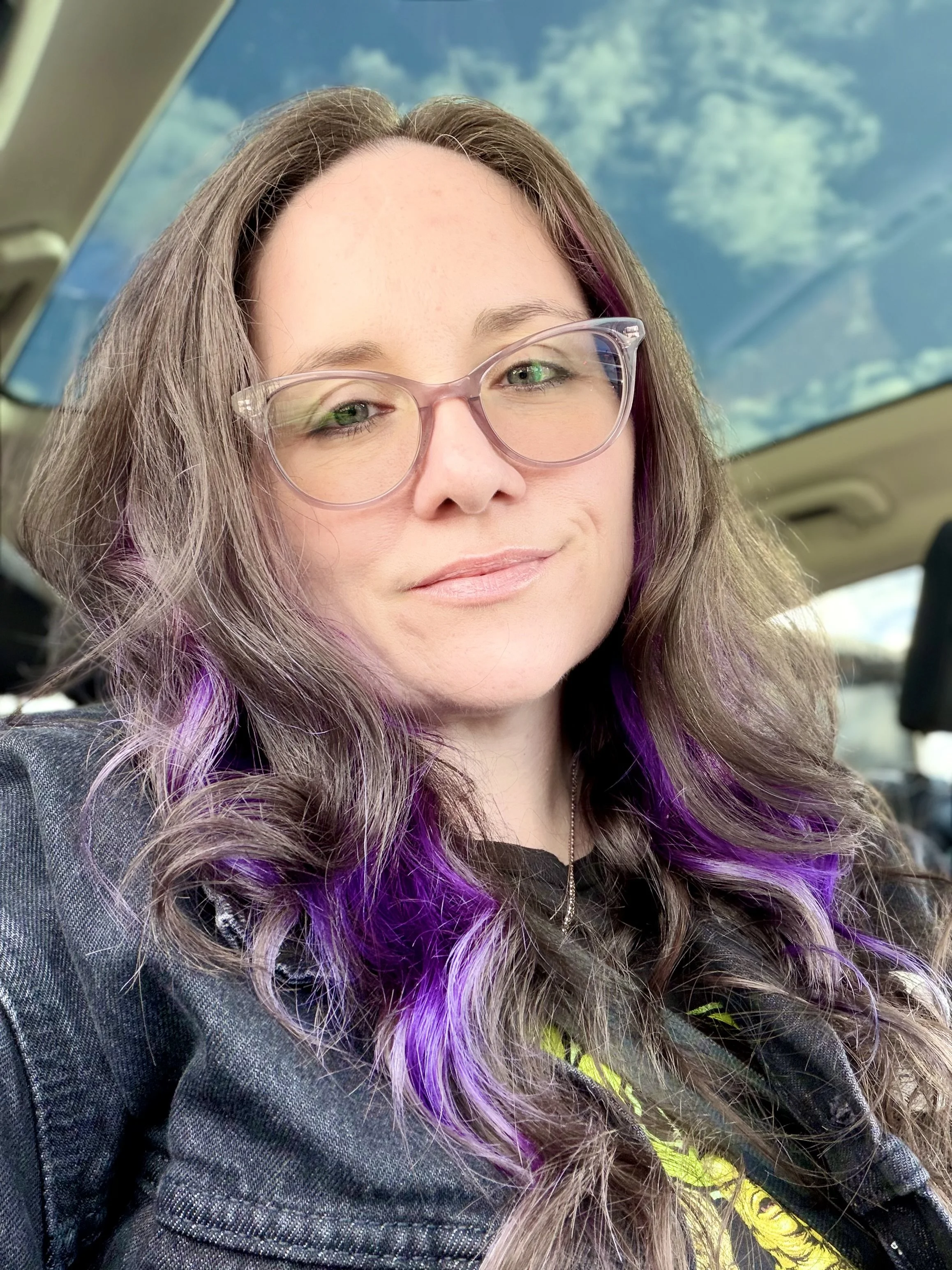 Kelly-Ann, a woman with wavy brown and purple hair, wearing glasses and a black denim jacket, is sitting inside a vehicle with a view of a blue sky and clouds through the sunroof and smiling at the camera.
