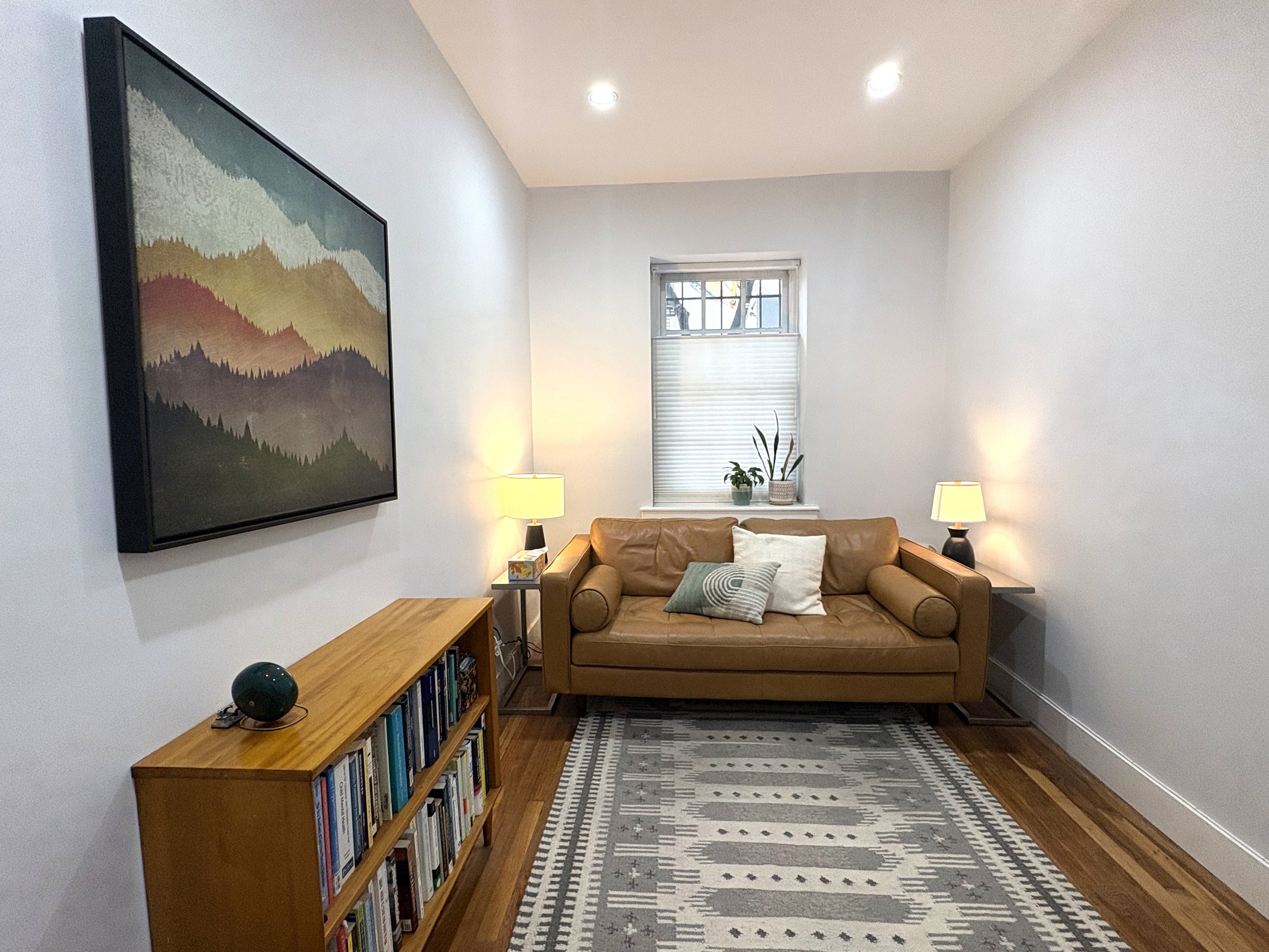 Therapy treatment room photo with a painting of a mountain landscape, a window with a white blind above a brown leather sofa, two table lamps on side tables, a wooden bookshelf filled with books, and a patterned rug on the hardwood floor.