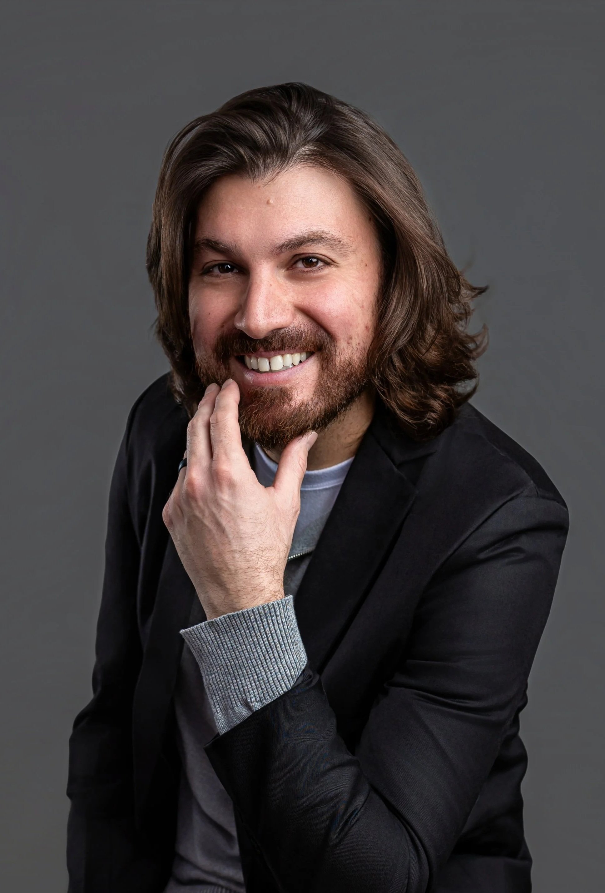A man with long brown hair and a beard smiling, wearing a black blazer and a gray sweater, posing against a gray background.