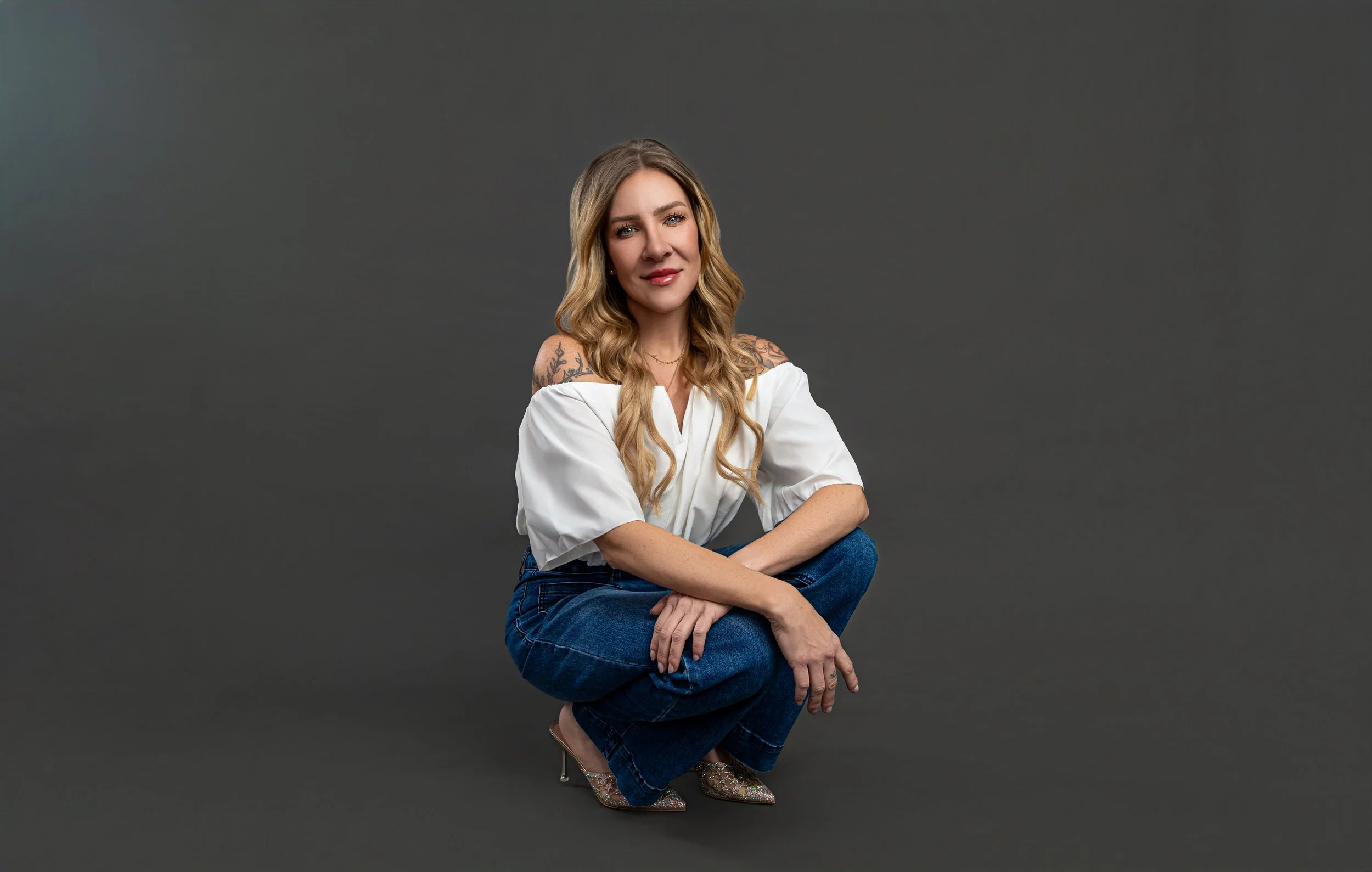 A woman with long wavy blonde hair and tattoos on her shoulders squats on a dark gray background, wearing a white off-the-shoulder top, blue jeans, and sparkly high heels.