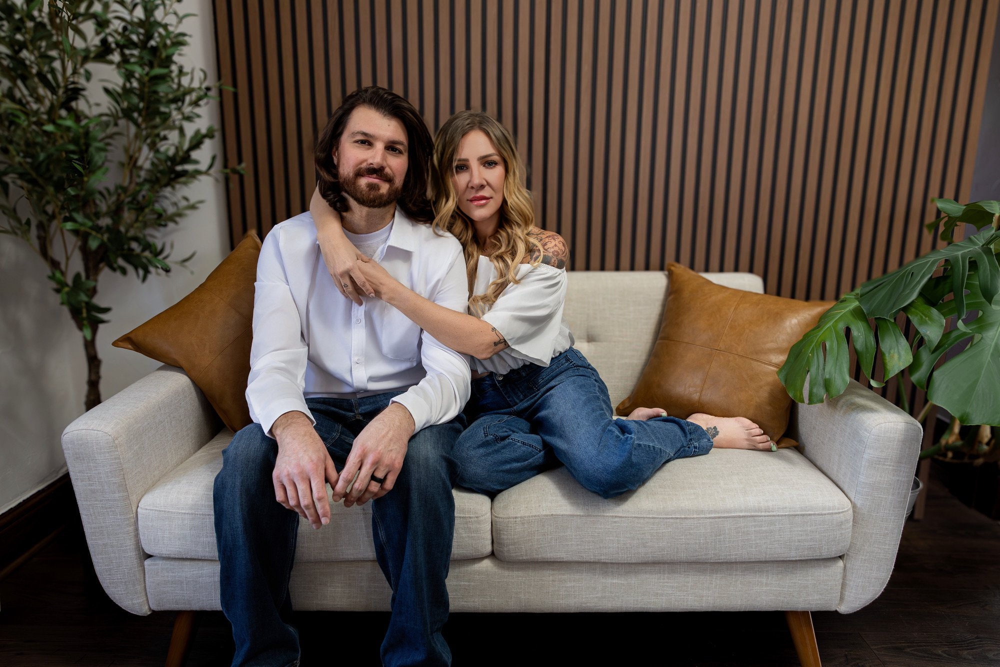 A man and woman sitting closely on a beige couch with brown pillows, surrounded by green plants, in a room with a wooden slat wall in the background.