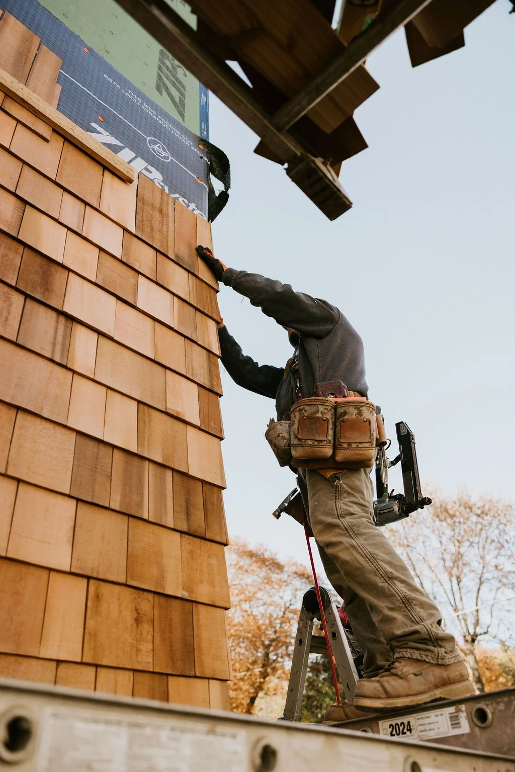 A worker on a ladder installing wooden shingles on the exterior of a building.