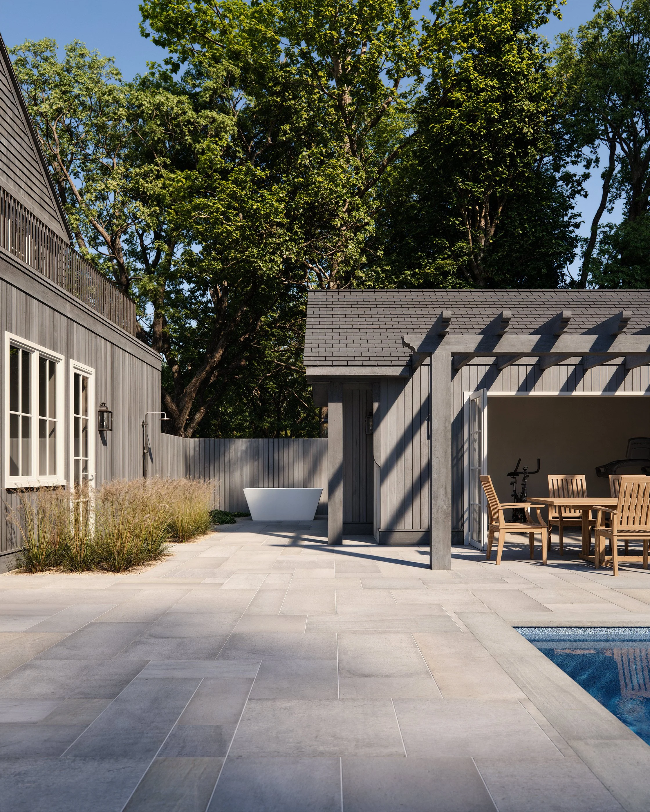 Outdoor patio with gray pavers, wooden furniture, pergola, and a pool next to a modern gray house with large trees in the background.