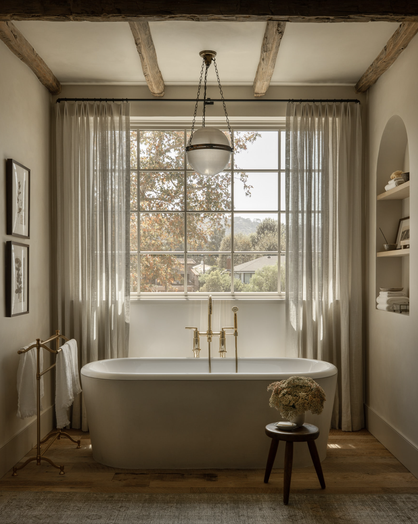 Elegant bathroom with a freestanding bathtub, large window with sheer curtains, wooden ceiling beams, and a pendant light. A stool with flowers is near the tub, and folded towels are on shelves.