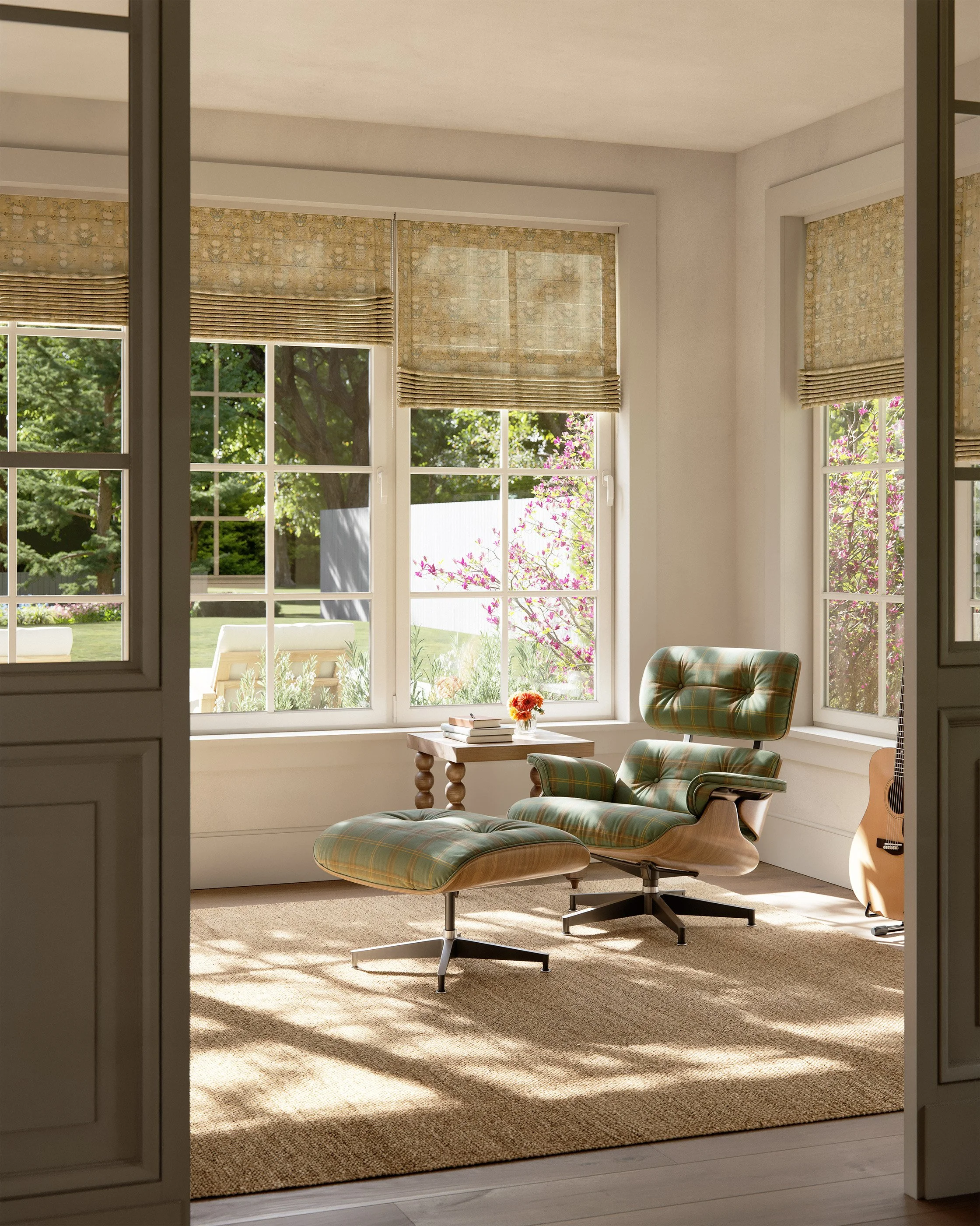 Bright sunlit room with large windows, plaid cushioned lounge chair, matching ottoman, small wooden side table with books and flowers, guitar leaning against the wall, and a beige rug on the floor.