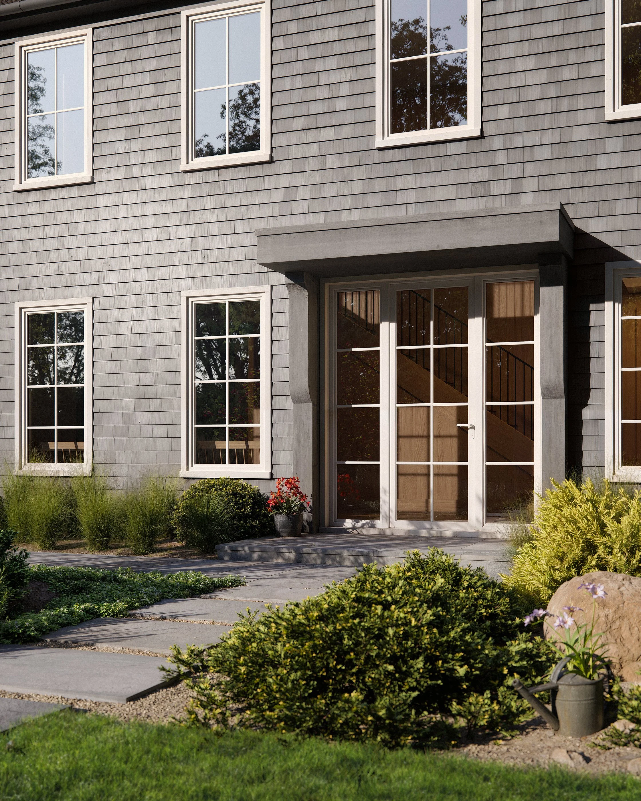 Front of a gray shingle-sided house with multiple white-framed windows, a glass door, and a landscaped garden. The garden features green shrubs, plants, and stepping stones leading to the entrance.
