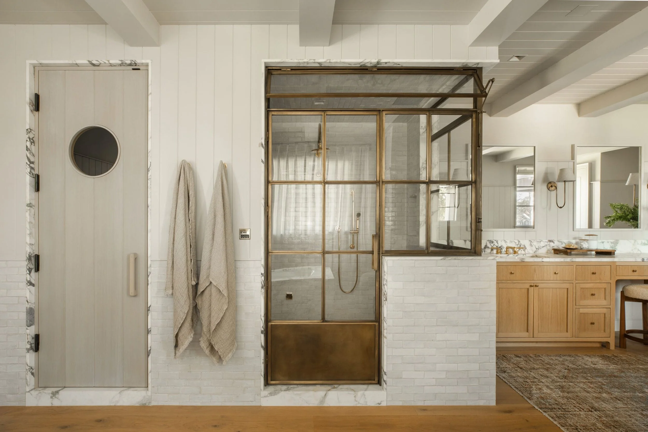Bathroom with a modern design featuring a glass and brass shower enclosure, two beige towels hanging on the wall, a wooden door with a circular window, and a wooden vanity with a marble countertop.