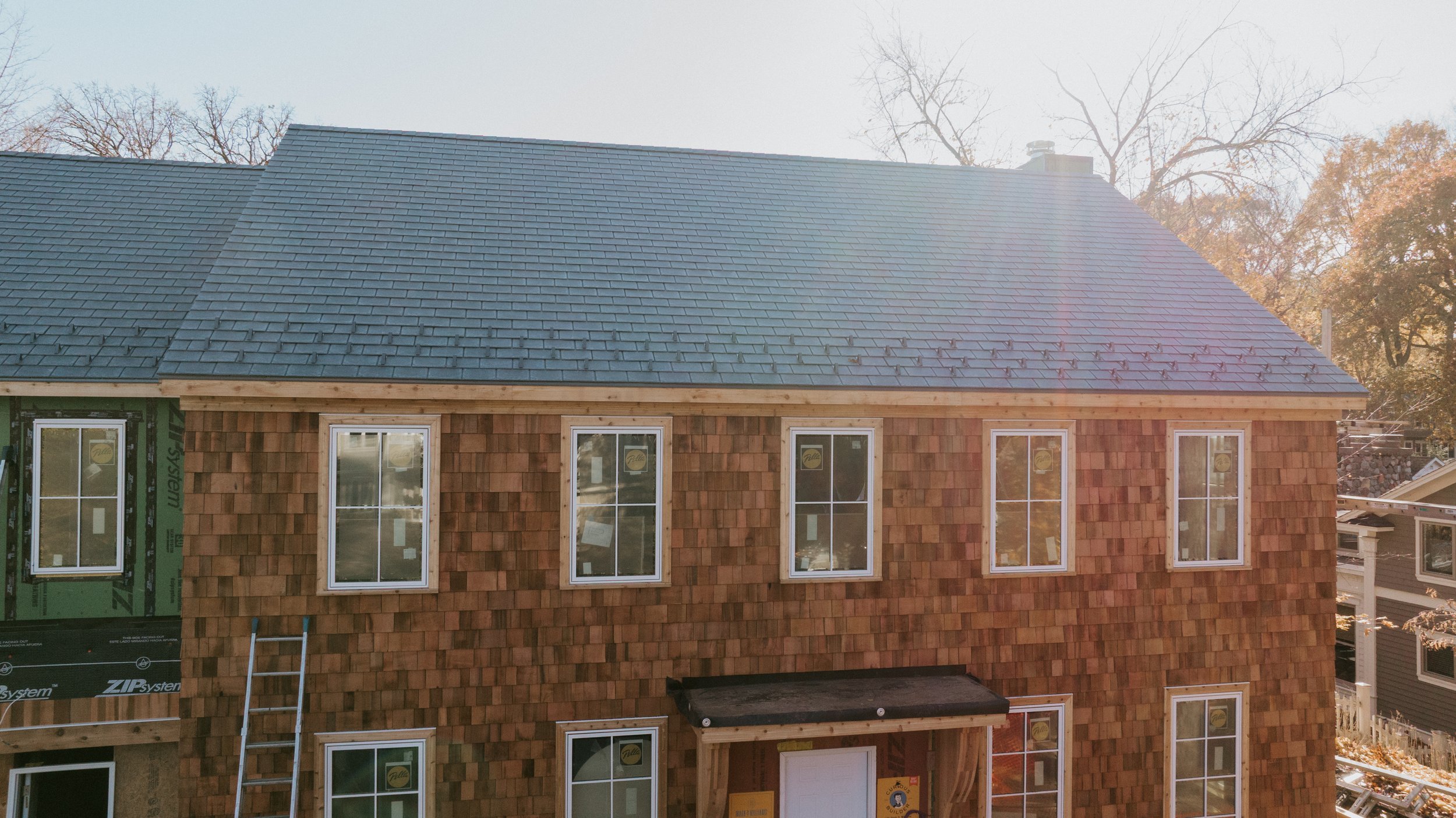 A newly constructed multi-story house with a gray shingled roof, wooden shingle siding, multiple windows, and a front door with a small covered porch, during daytime with bare trees in the background.