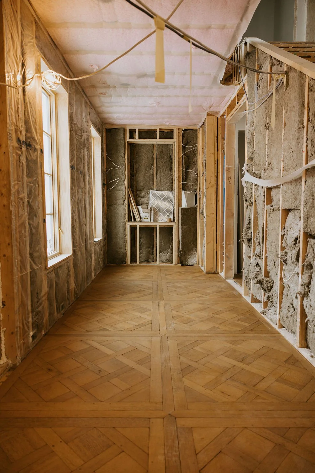 Room under renovation with exposed wall studs, insulation, wiring, and newly installed wooden floor.