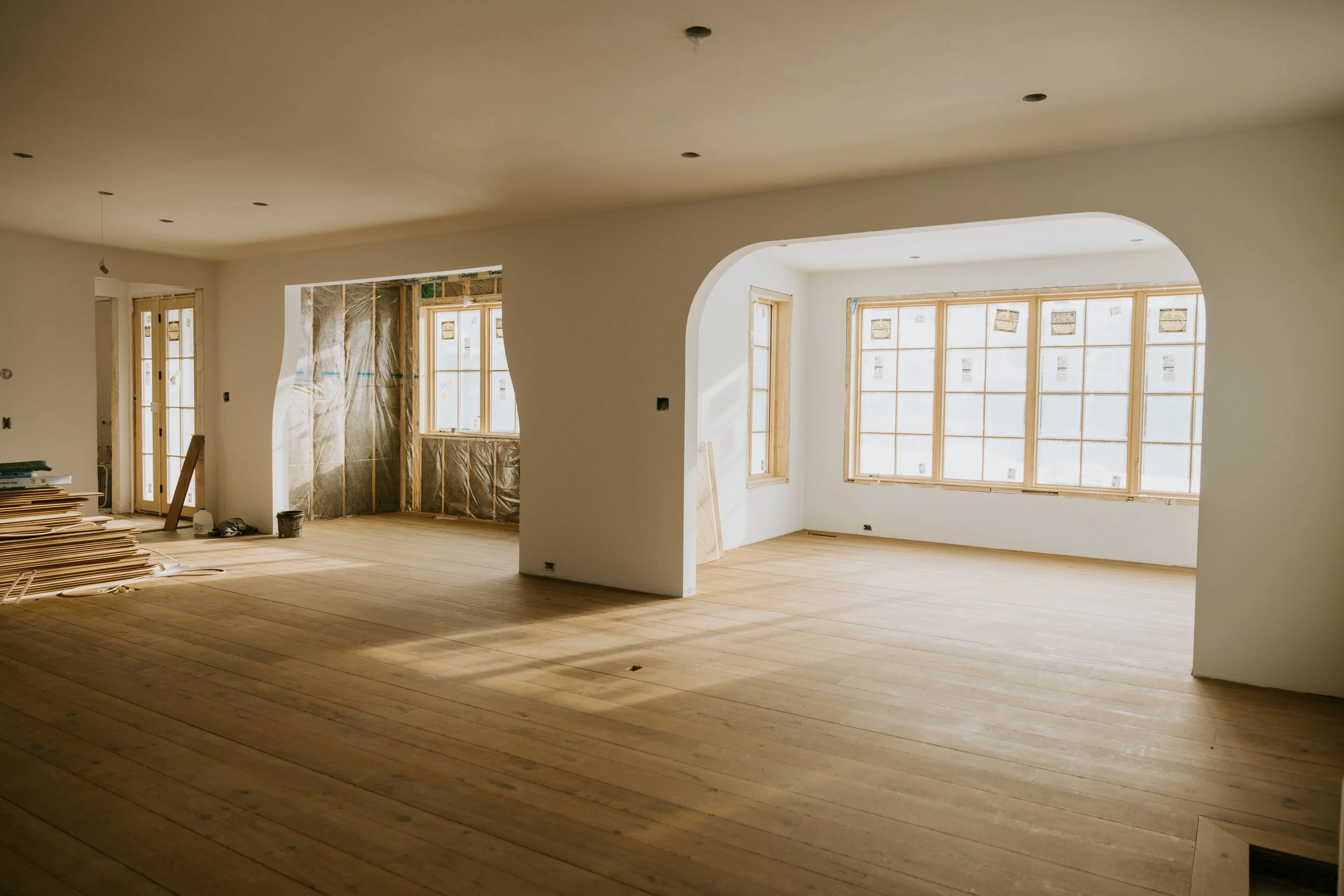 An empty room in a house under construction with unfinished walls, large windows, and wooden flooring.