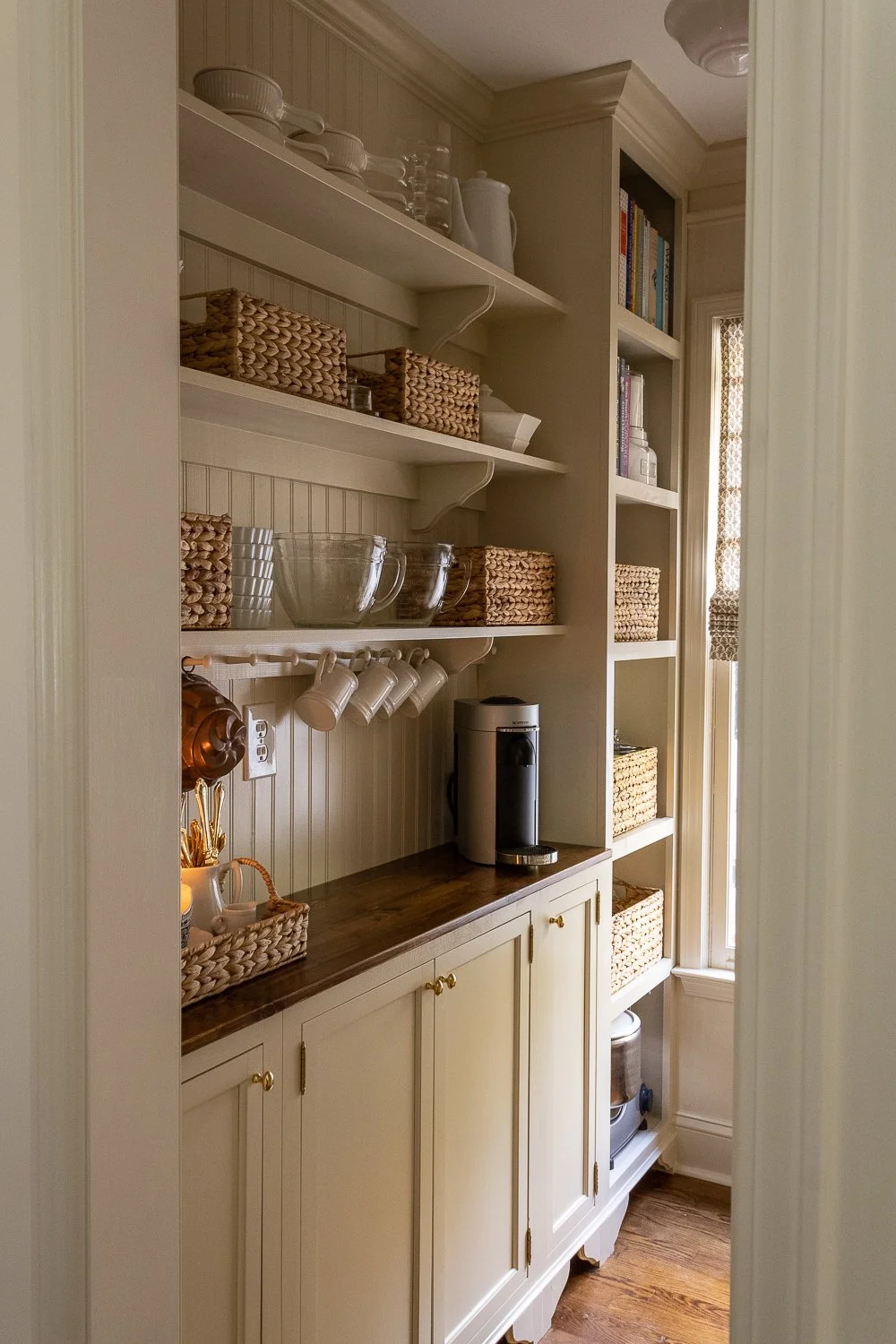 Organized pantry with wooden shelves, woven baskets, dishware, coffee maker, and books.