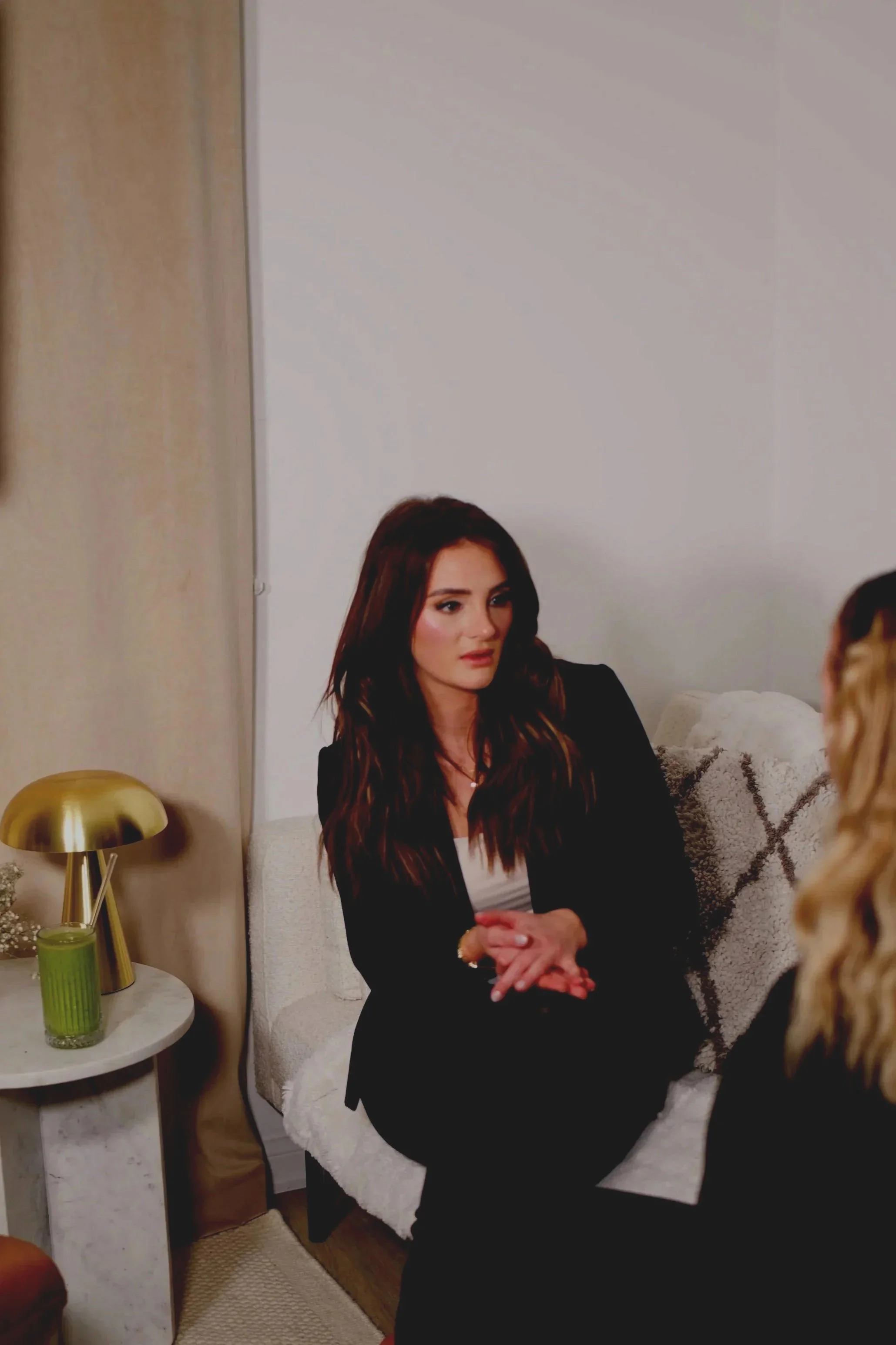 A woman with long brown hair wearing a black blazer and light-colored blouse, sitting on a white sofa with her hands clasped, engaging with someone not fully visible in the frame. There is a small round side table next to her with a gold table lamp and a green candle, in a cozy living room setting.