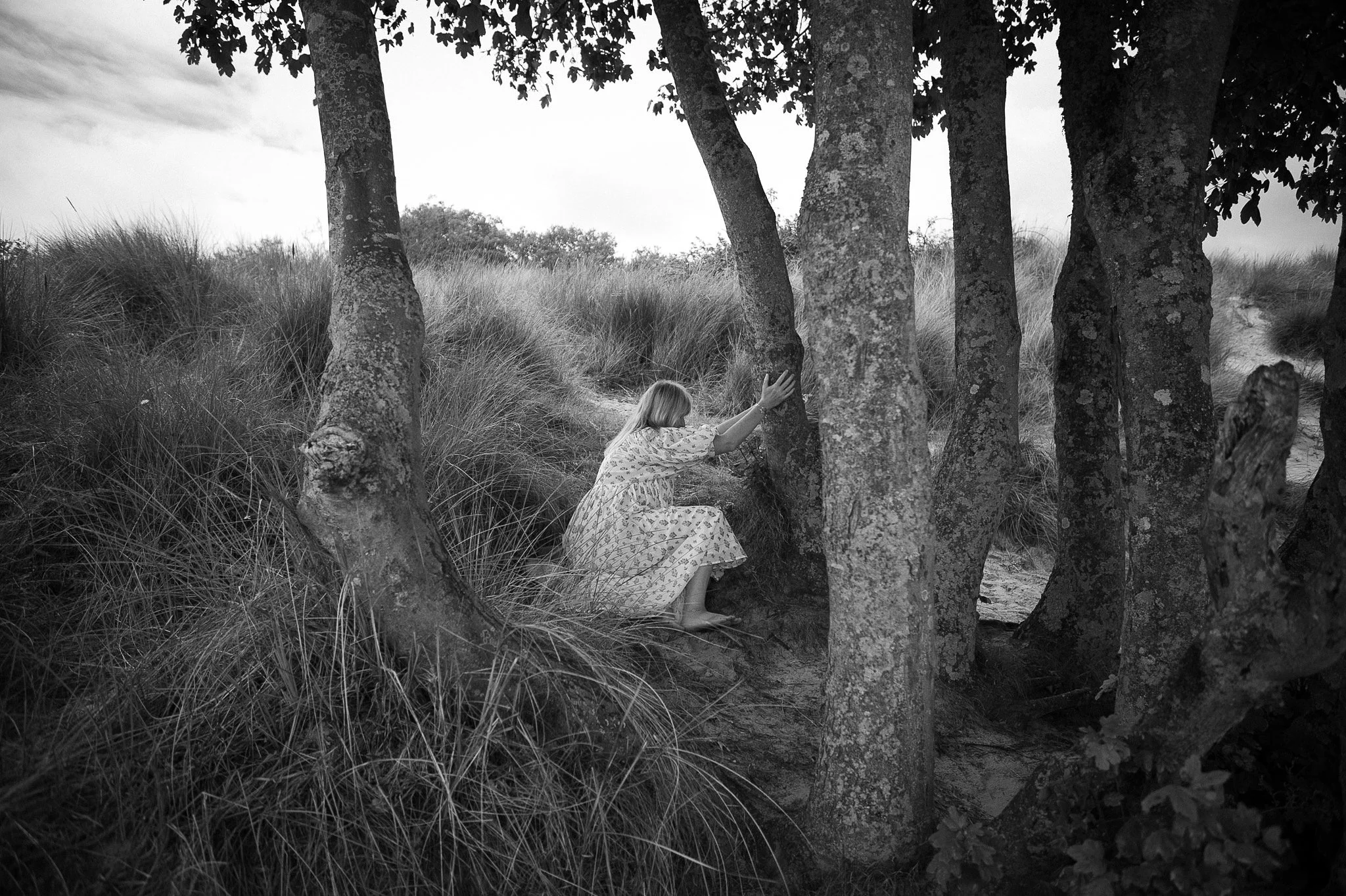 Black and white photo of a woman in a floral dress crouching among trees in a grassy area.