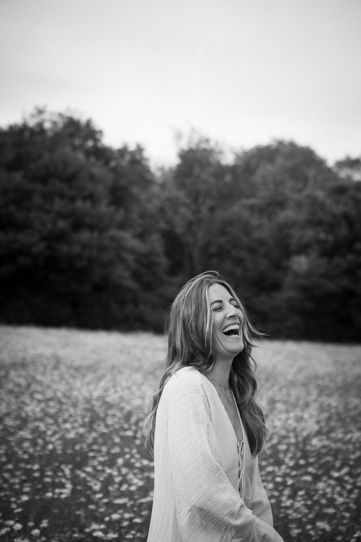 A woman laughing outdoors in a field of flowers with trees in the background, in black and white. Soulful branding photo shoot in Kent, England.