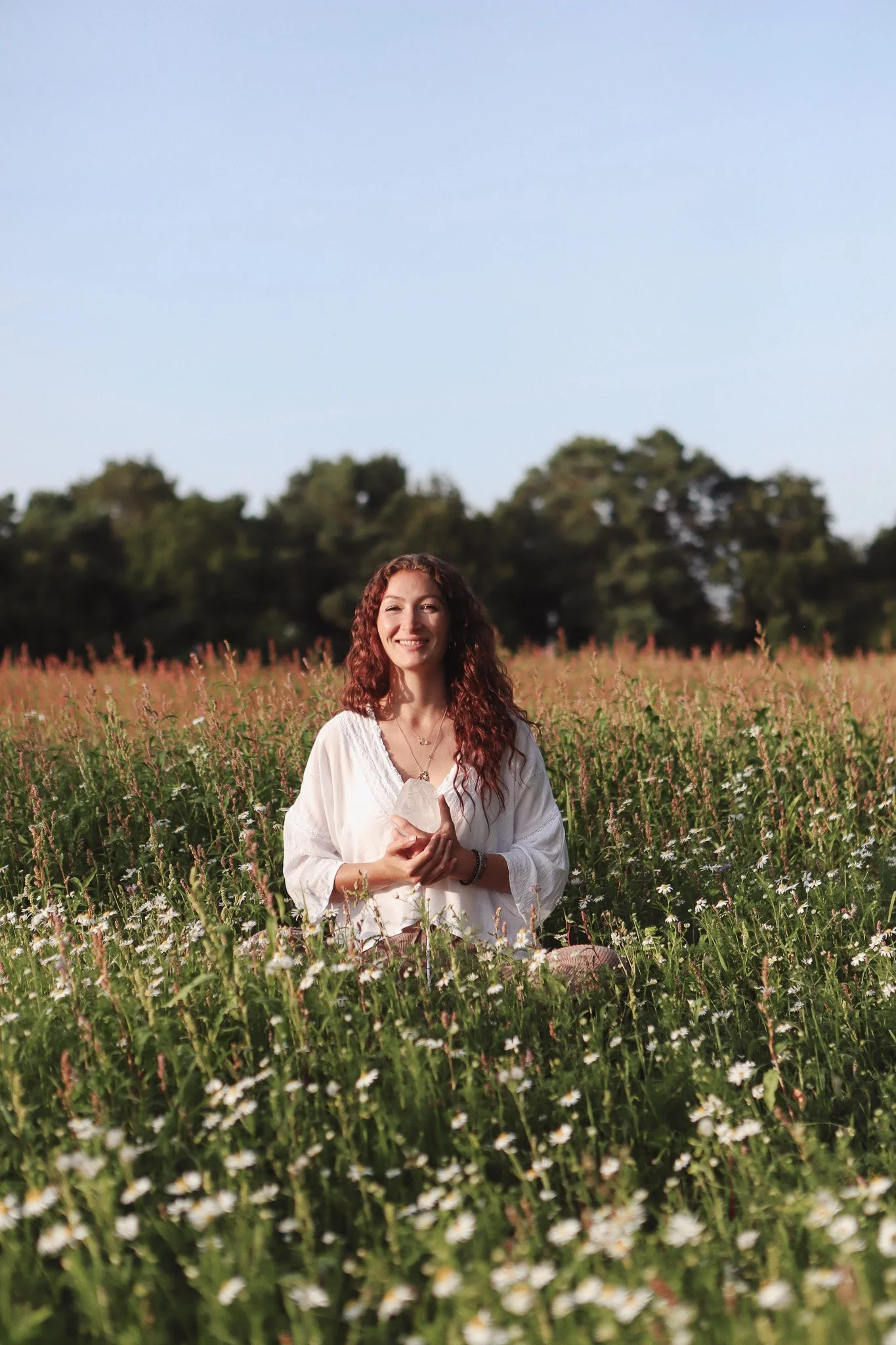 Woman in white dress sitting in a field of daisies, holding a large crystal, with trees in the background.