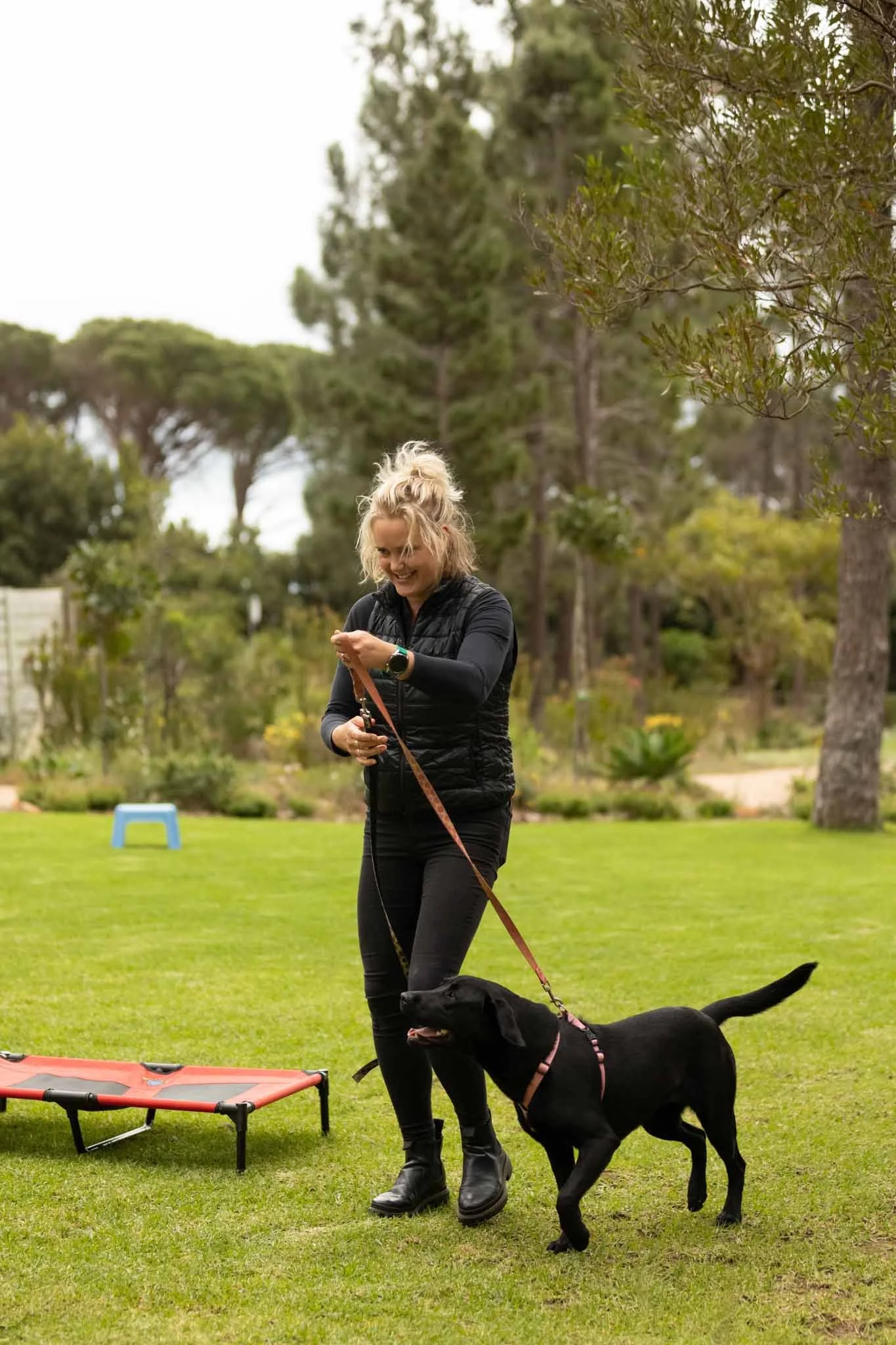 A woman with blonde hair in a ponytail smiling as she holds a leash with a black dog on a grassy field with trees in the background.
