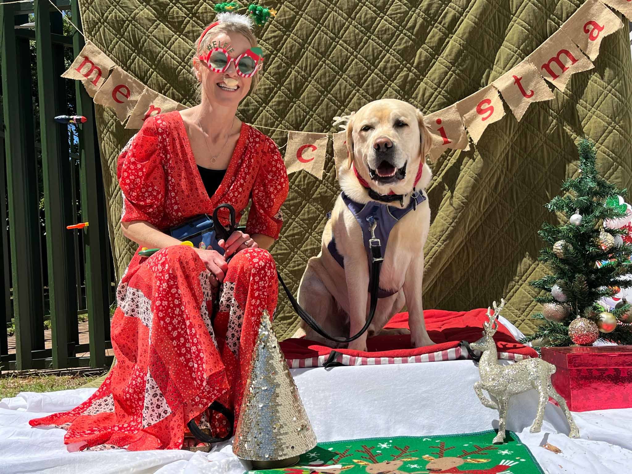 A woman dressed in red with holiday glasses and a Santa hat poses with a smiling yellow Labrador retriever wearing a blue harness. They are outdoors with Christmas decorations including a small decorated tree, reindeer figures, and a festive rug, wit