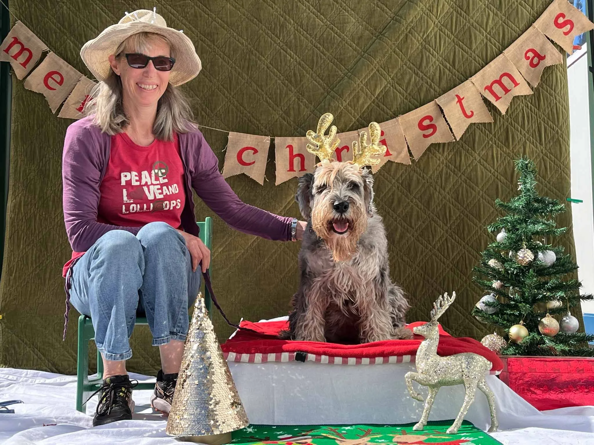 A woman sitting on a green chair with a dog in a holiday setting. The woman wears a wide-brimmed hat, sunglasses, a red shirt, and a purple jacket. The dog, seated on a red mat, has reindeer antlers. In the background, a green quilted backdrop has a 