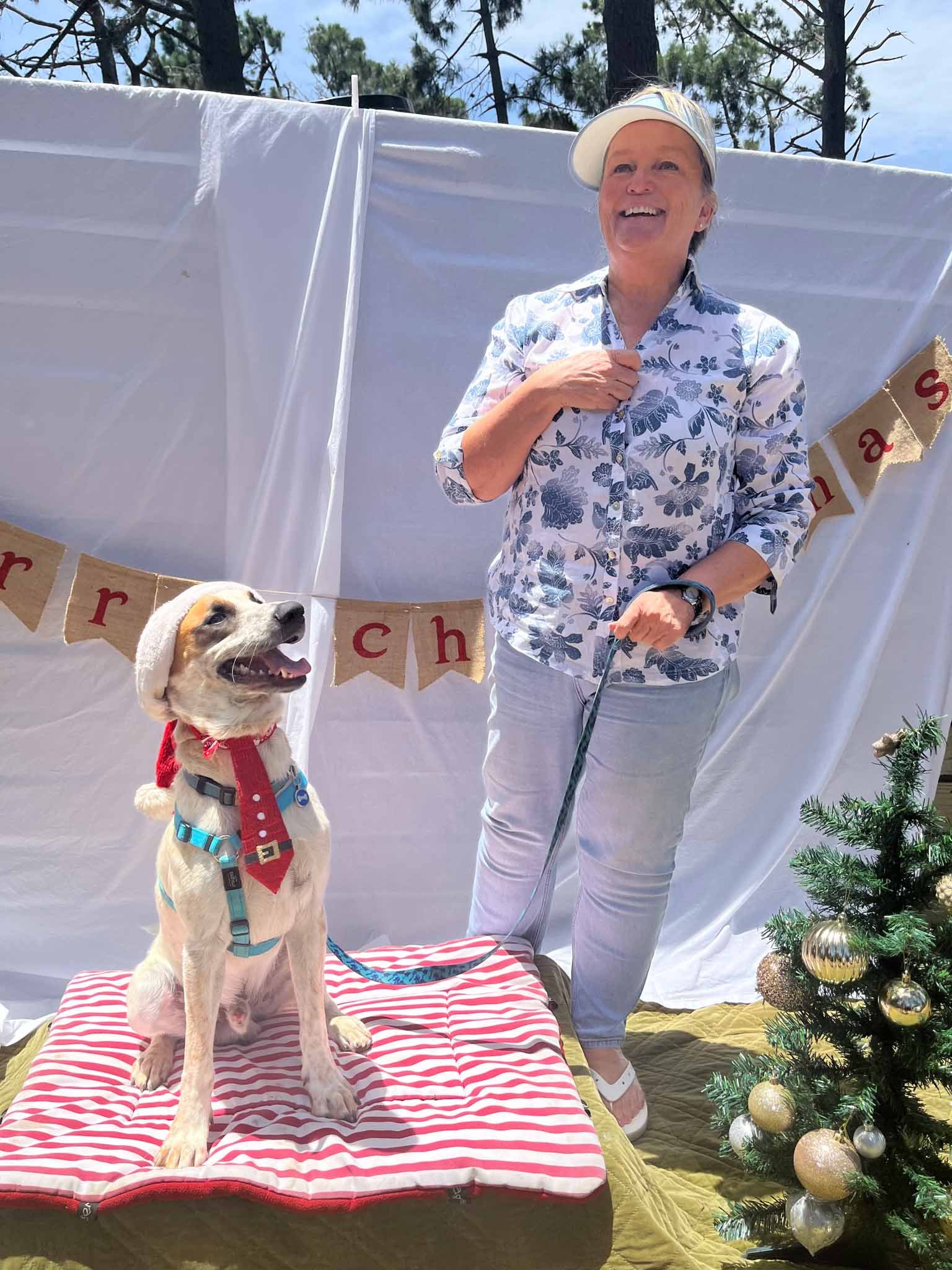 A woman and a puppy pose outside during a Christmas celebration. The woman is smiling, wearing a white cap and a floral shirt. The puppy, wearing a red and white outfit, sits on a striped cushion. There is a Christmas tree and a banner that says 'Mer