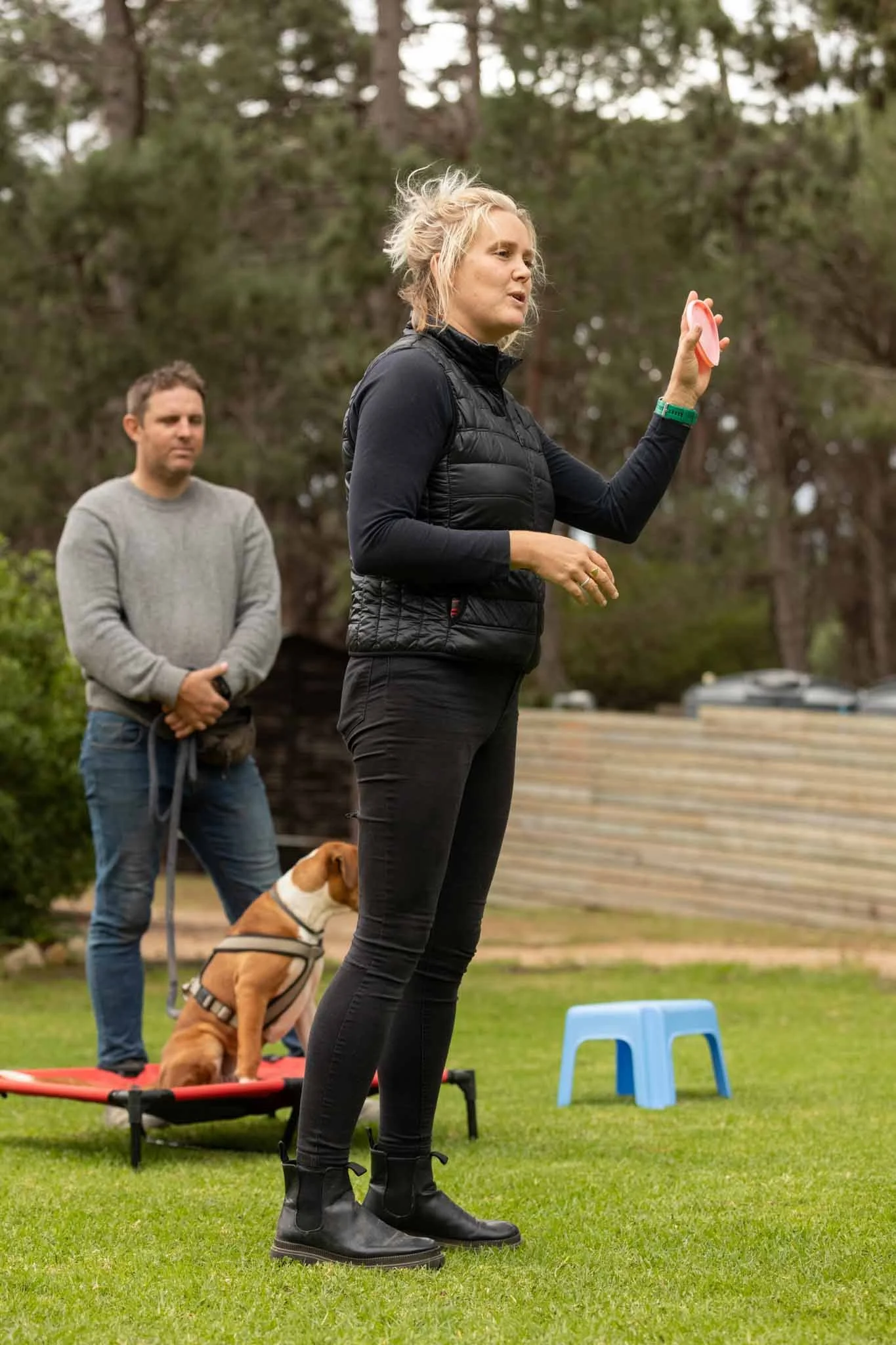 A woman instructing in a park with a dog on a platform and a man standing behind her, observing.