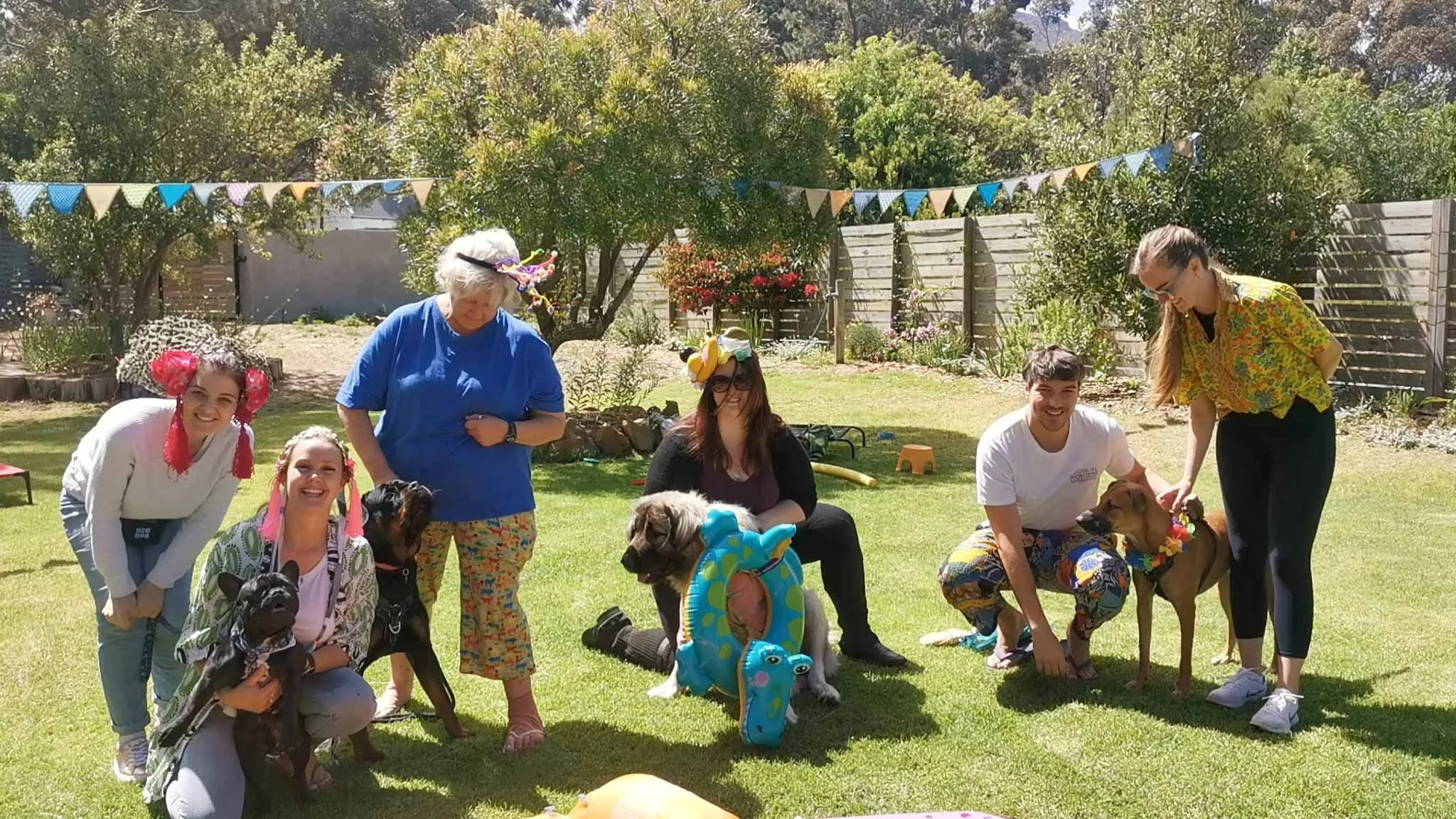 Group of people and dogs celebrating in a backyard, some wearing party accessories, with bunting and trees in the background.