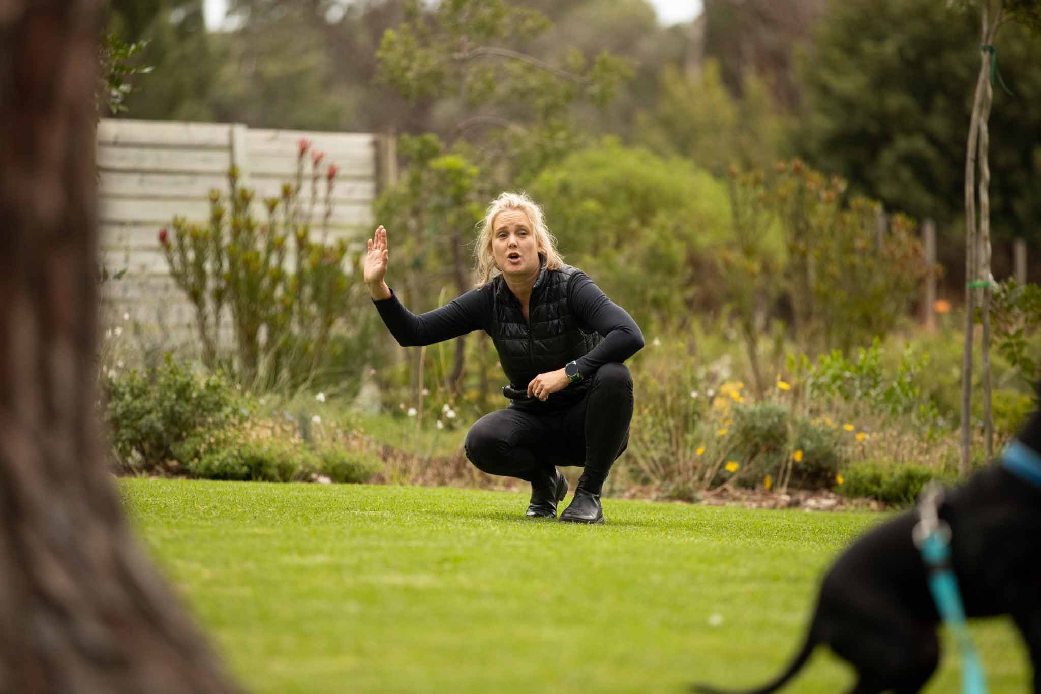 A woman crouching on a lawn in an outdoor garden, gesturing with her right hand while talking or explaining something, with trees and a fence in the background.