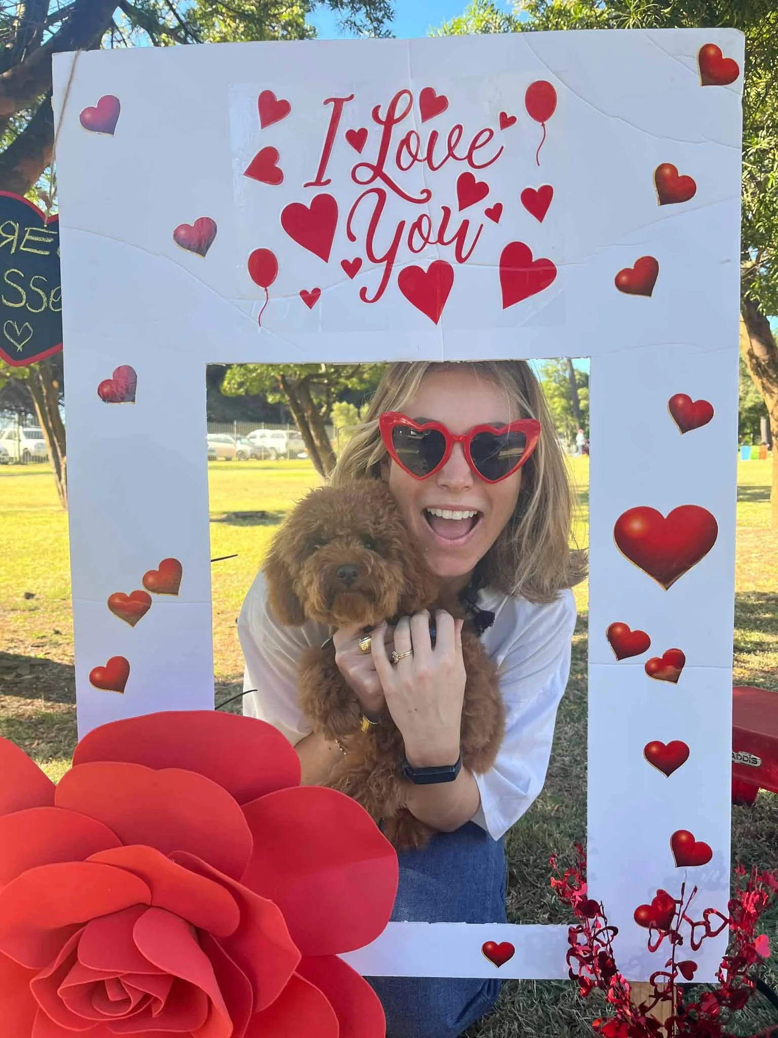 A woman with blonde hair wearing red heart-shaped sunglasses, smiling and holding a small brown poodle, posing behind a large white Valentine-themed photo frame decorated with red hearts and the words 'I Love You' in red. There are large red paper ro
