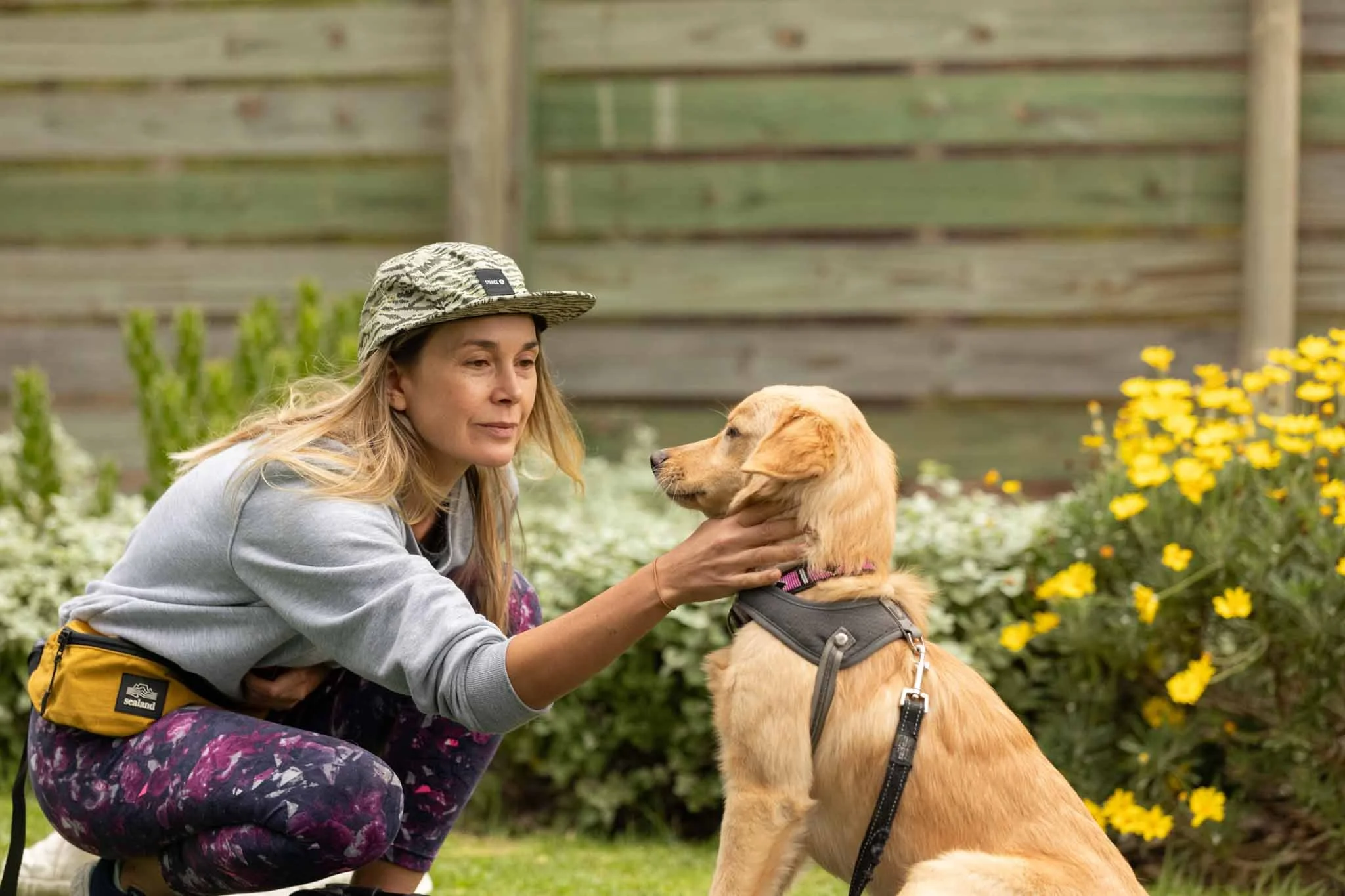 A woman kneeling and gently holding a yellow Labrador Retriever's face in her hands in a garden with yellow flowers and a wooden fence in the background.