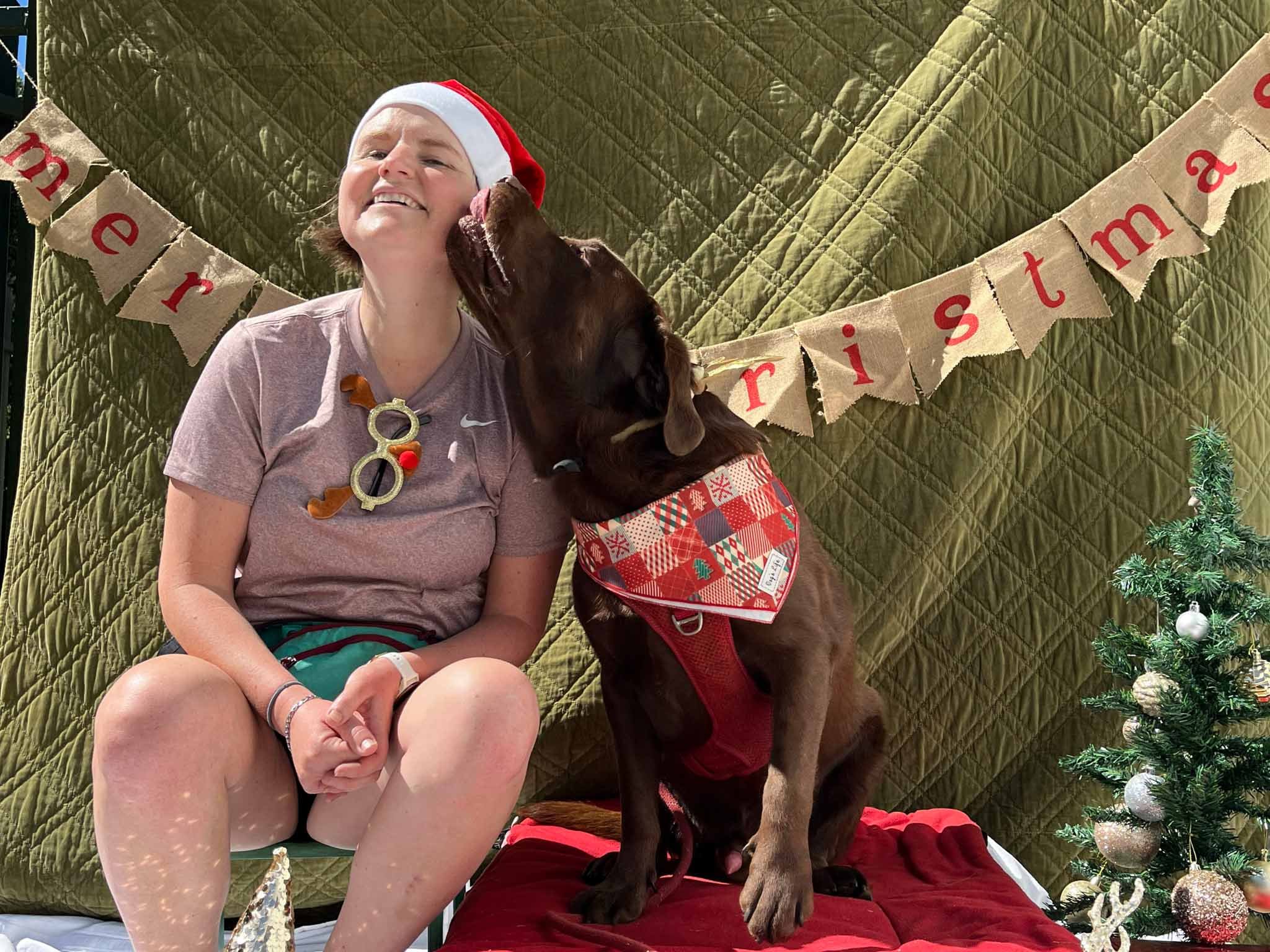 A young woman wearing a Santa hat sitting on a stool, smiling as a brown dog wearing a red and white bandana kisses her cheek on a red blanket. They are surrounded by Christmas decorations including a small decorated Christmas tree and a festive bann