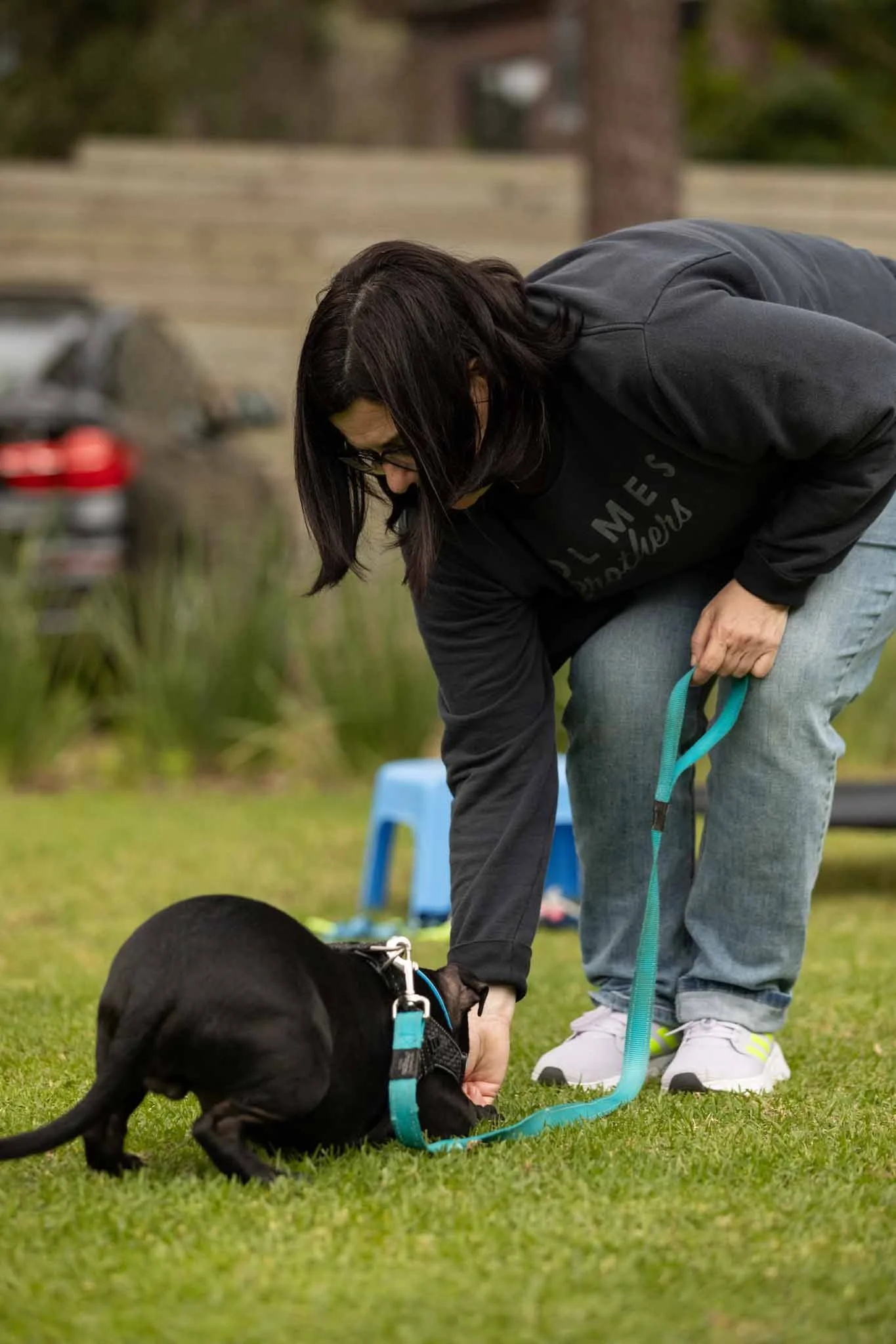 A woman in eyeglasses crouches down and holds the leash of a black puppy in a grassy outdoor area, with a wooden fence, trees, a small blue stool, and a parked car in the background.