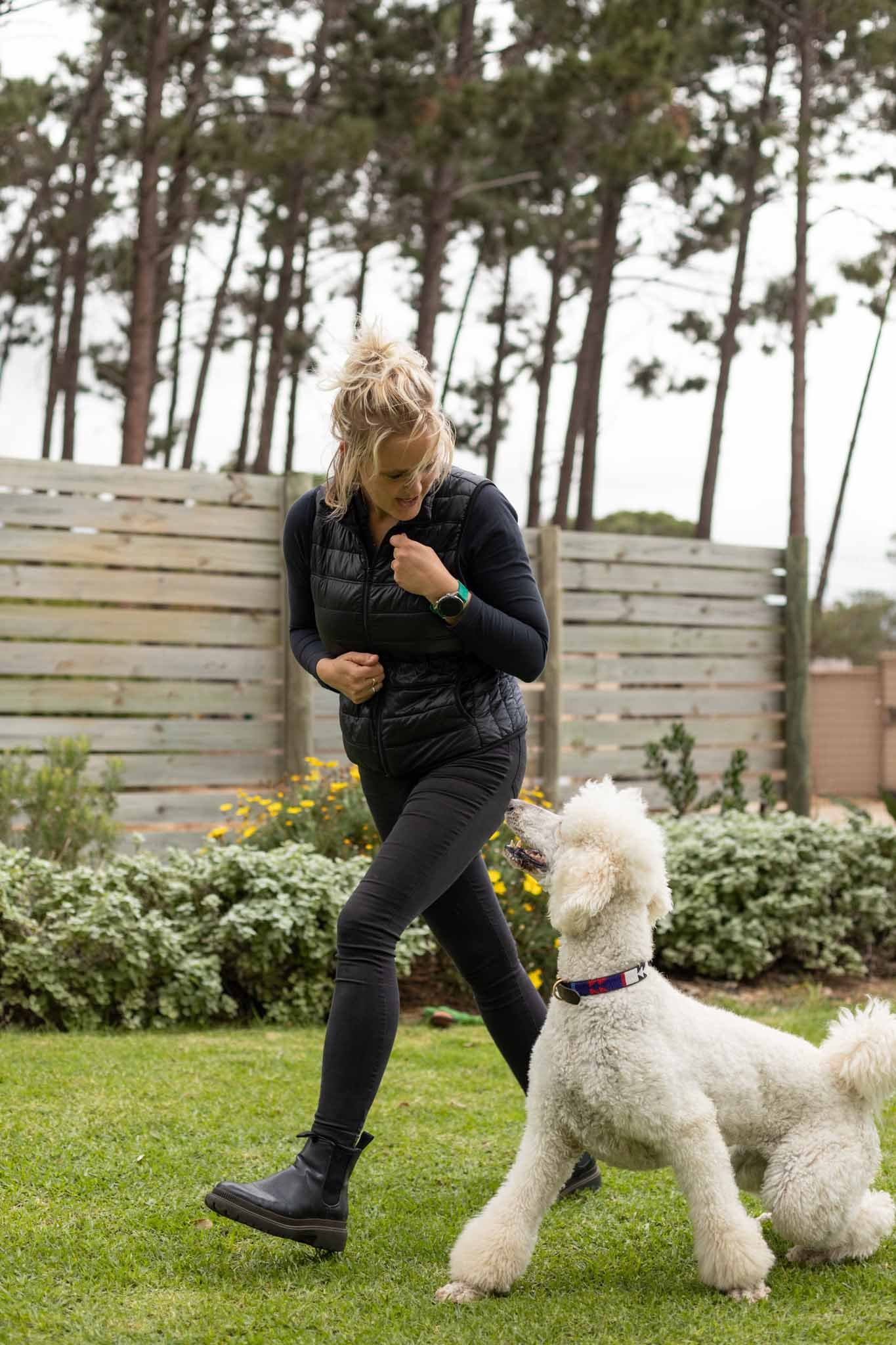 A woman playing with a large, fluffy white dog in a backyard with a green lawn and a wooden fence, surrounded by trees and bushes.