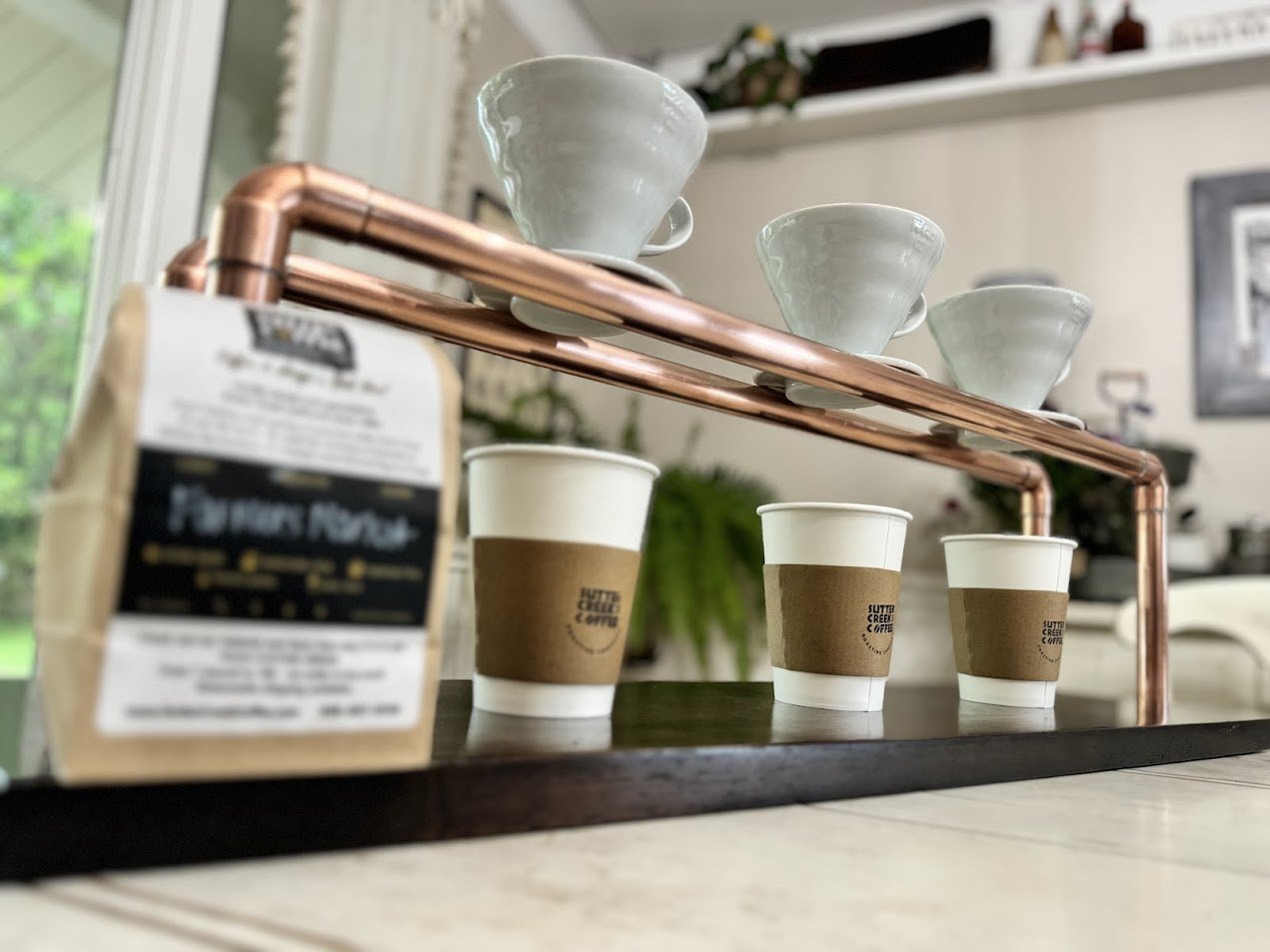 Three coffee cups with brown sleeves and a bag of Coffee Beans on a black tray with a copper handle, indoors with kitchen decor in the background.
