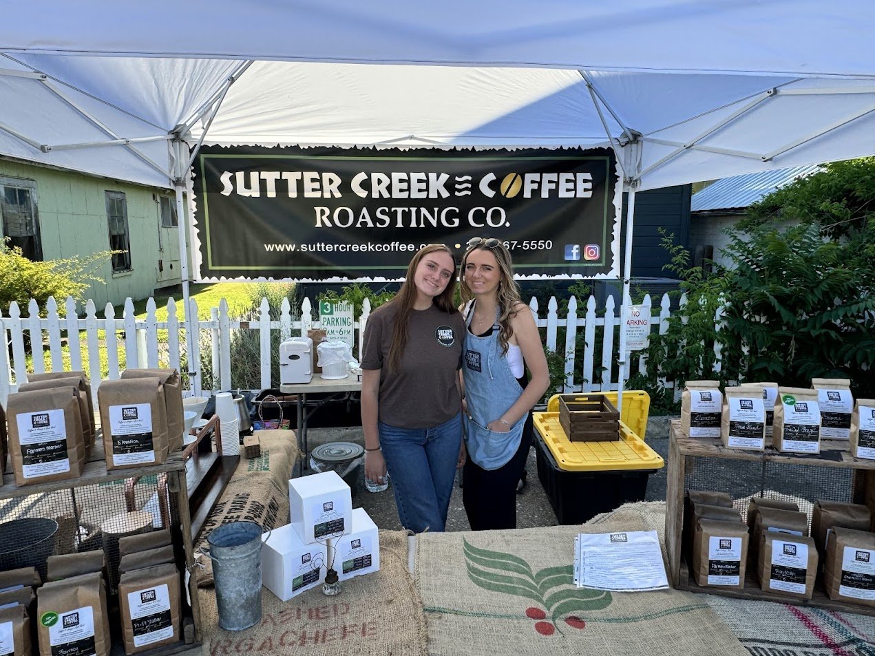 Two women smiling at a coffee booth with Sutter Creek Coffee signs, bags of coffee on tables, and a white picket fence in the background.