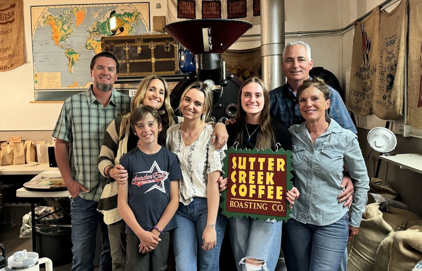 Group of seven people standing inside a coffee roasting shop, smiling, with a coffee roaster in the background and a sign that reads 'Sutter Creek Coffee Roasting Co.'