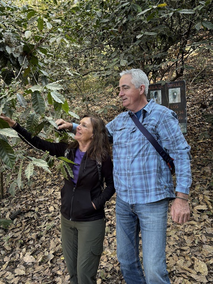 A man and woman exploring a dense forest area with large green leaves, and a sign in the background in Spanish.
