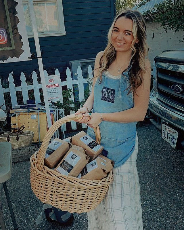A smiling woman in a denim apron holding a wicker basket filled with brown paper packages, standing outside near a black vehicle and a white picket fence.