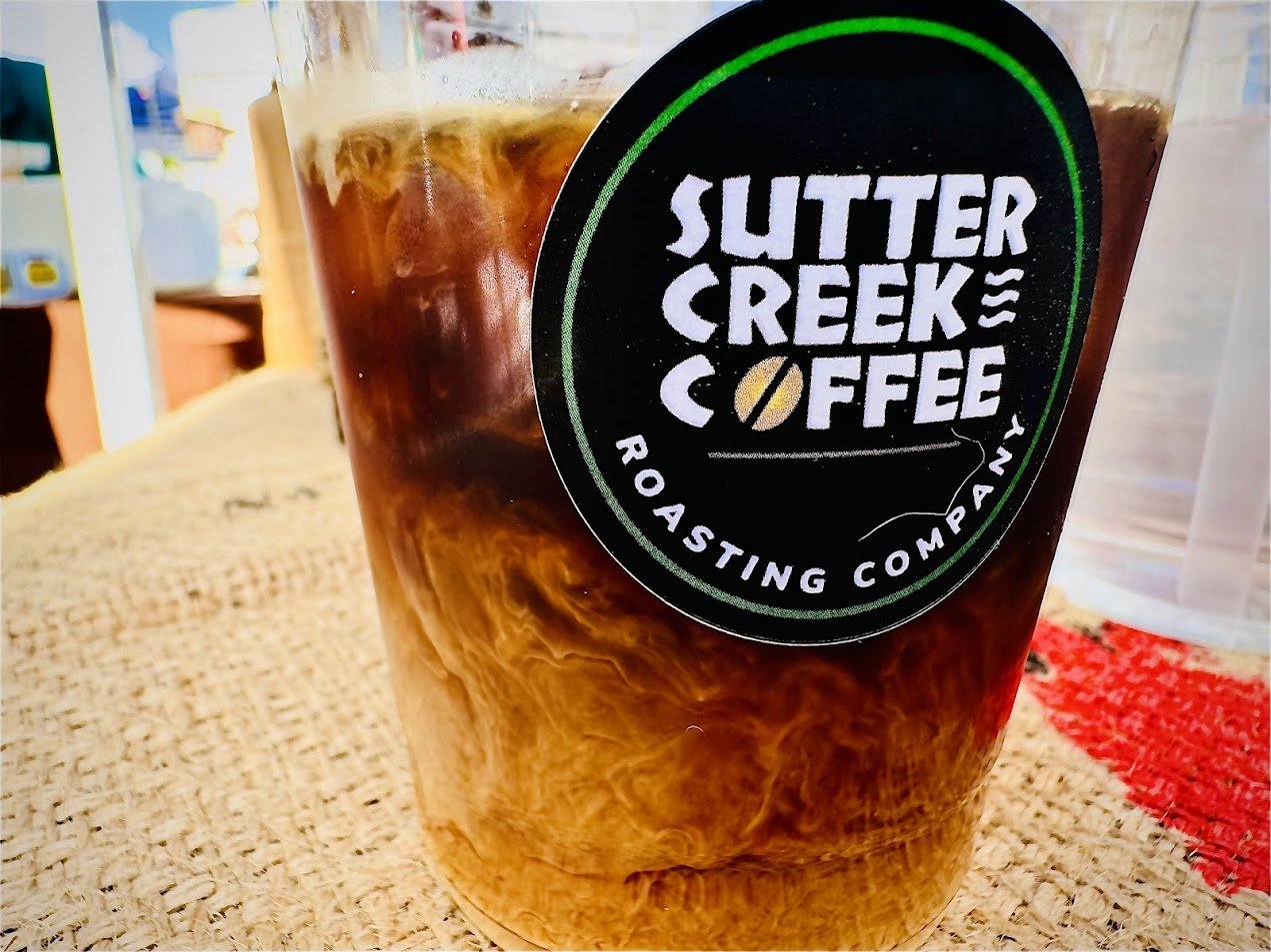 A plastic cup of iced coffee with a 'Sutter Creek Coffee Roasting Company' logo on it, placed on a woven surface with a beige cloth and a red cloth nearby.