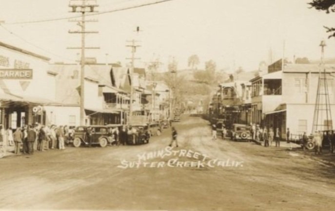 A black and white photo of a small town street scene with vintage cars parked along the sides, people walking on the sidewalk, and older buildings lining the street. Text at the bottom of the photo reads, 'Main Street Sutter Creek, Calif.'