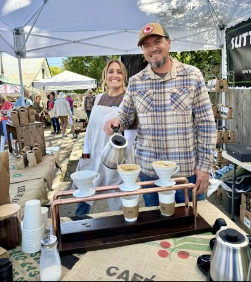A man and woman smiling at an outdoor coffee stand with cups of brewed coffee and pour-over coffee makers on display.
